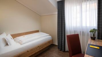 Habitación de hotel con una cama de madera, una silla roja, una mesita con una planta y cortinas traslúcidas.