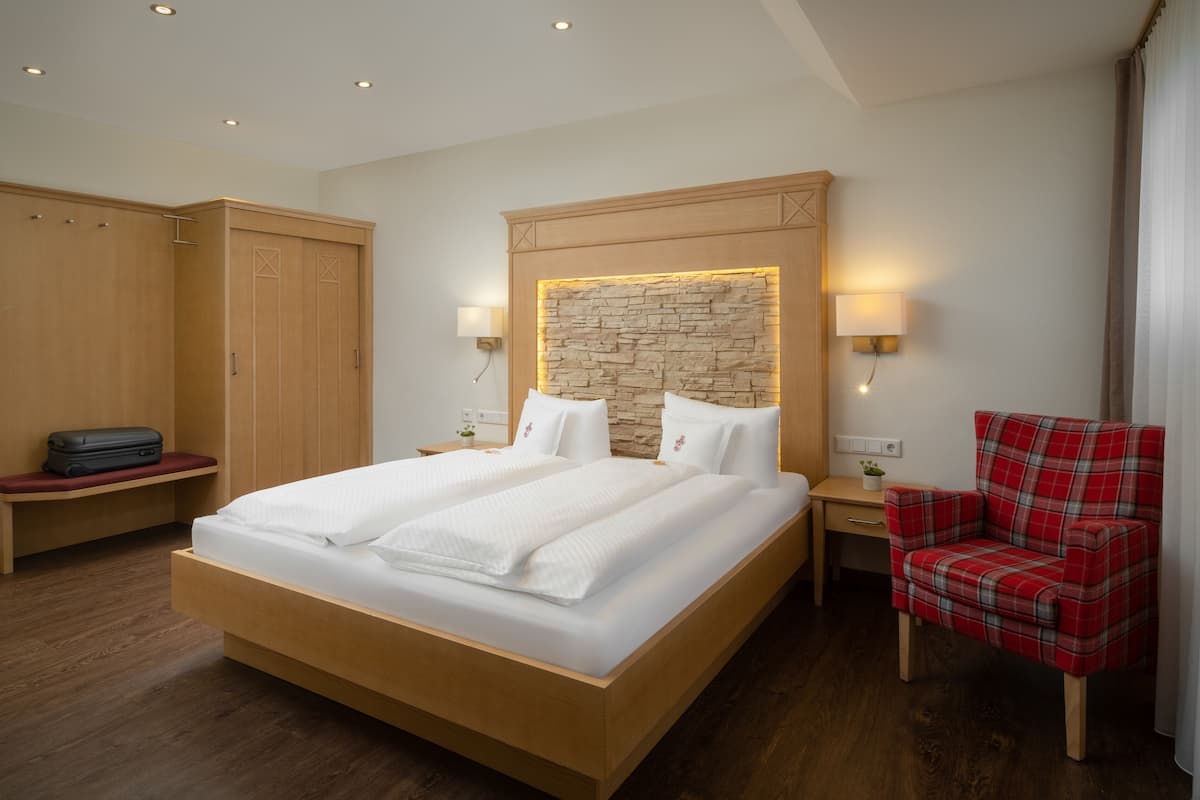Habitación de hotel con una cama grande, armario de madera, una silla de tela escocesa roja y una pared decorativa de piedra.