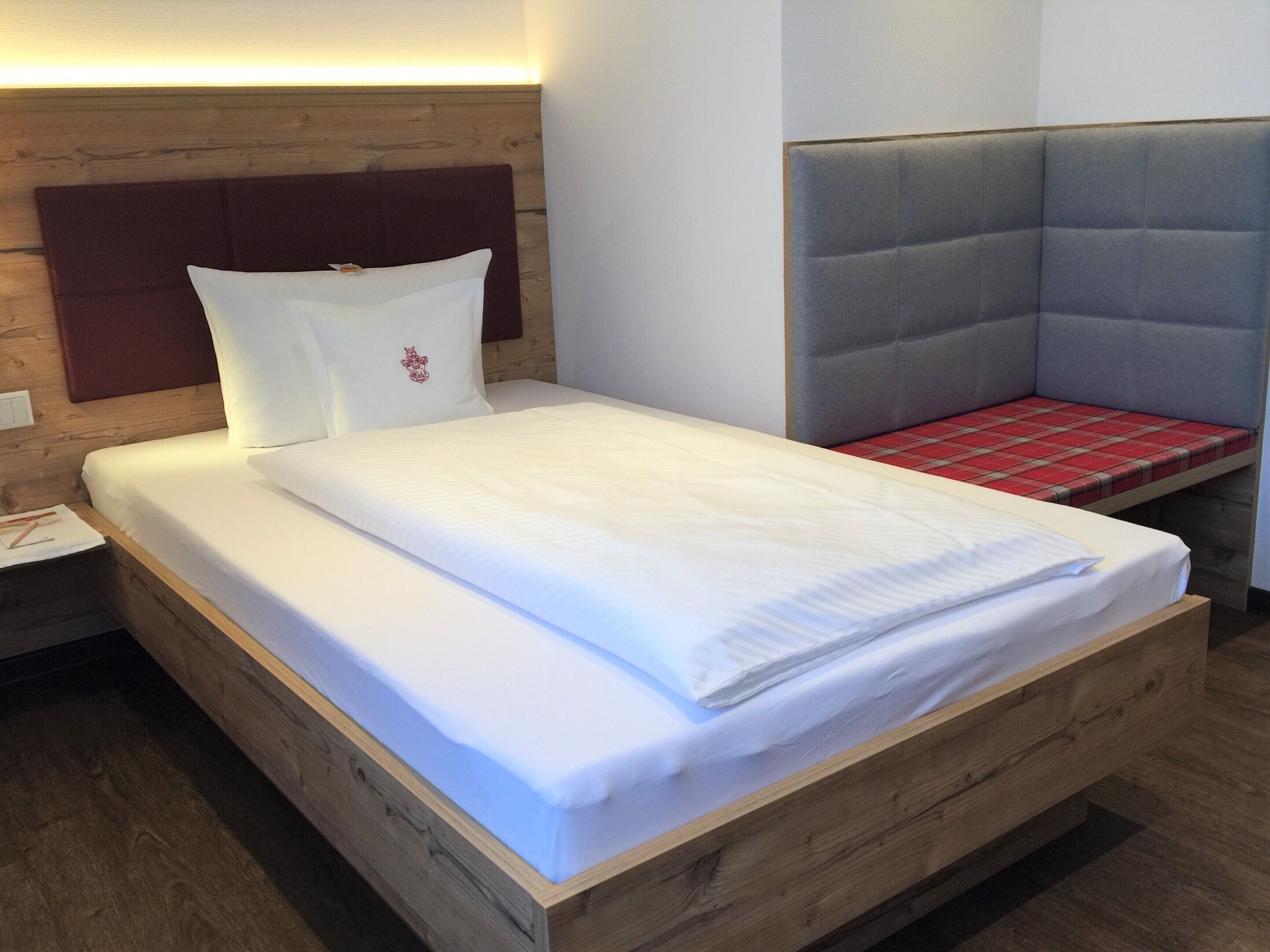 A neatly made bed with white linens, a wooden headboard, and a red plaid bench.