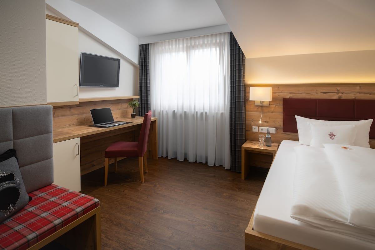 Habitación de hotel con cama, escritorio, laptop, silla, televisor y ventana con cortinas.