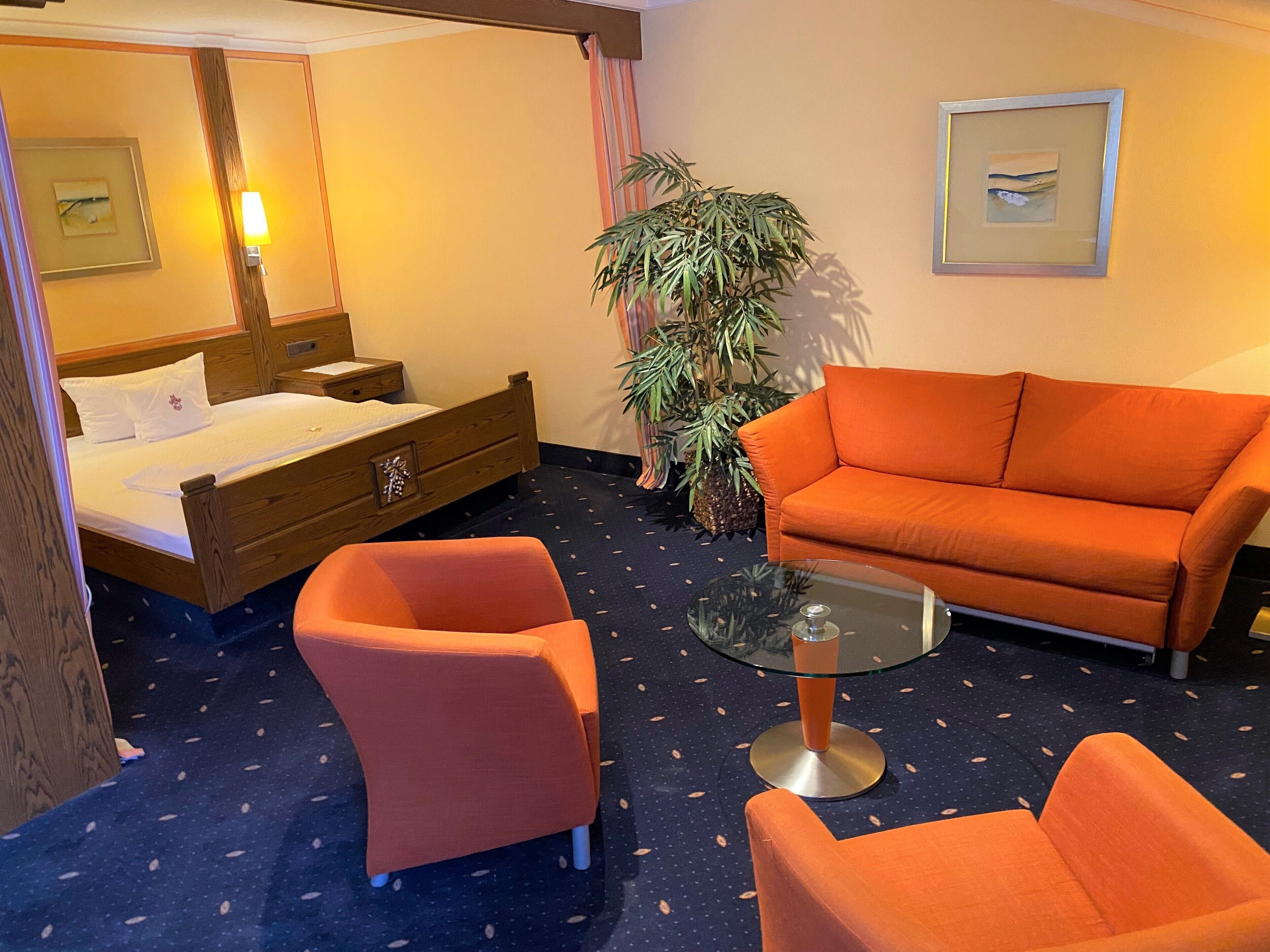 Habitación de hotel con una cama, dos sillones anaranjados, una mesa de centro de vidrio, una planta grande en maceta y cuadros en las paredes.