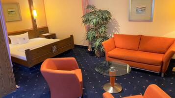 Habitación de hotel con una cama, dos sillones anaranjados, una mesa de centro de vidrio, una planta grande en maceta y cuadros en las paredes.
