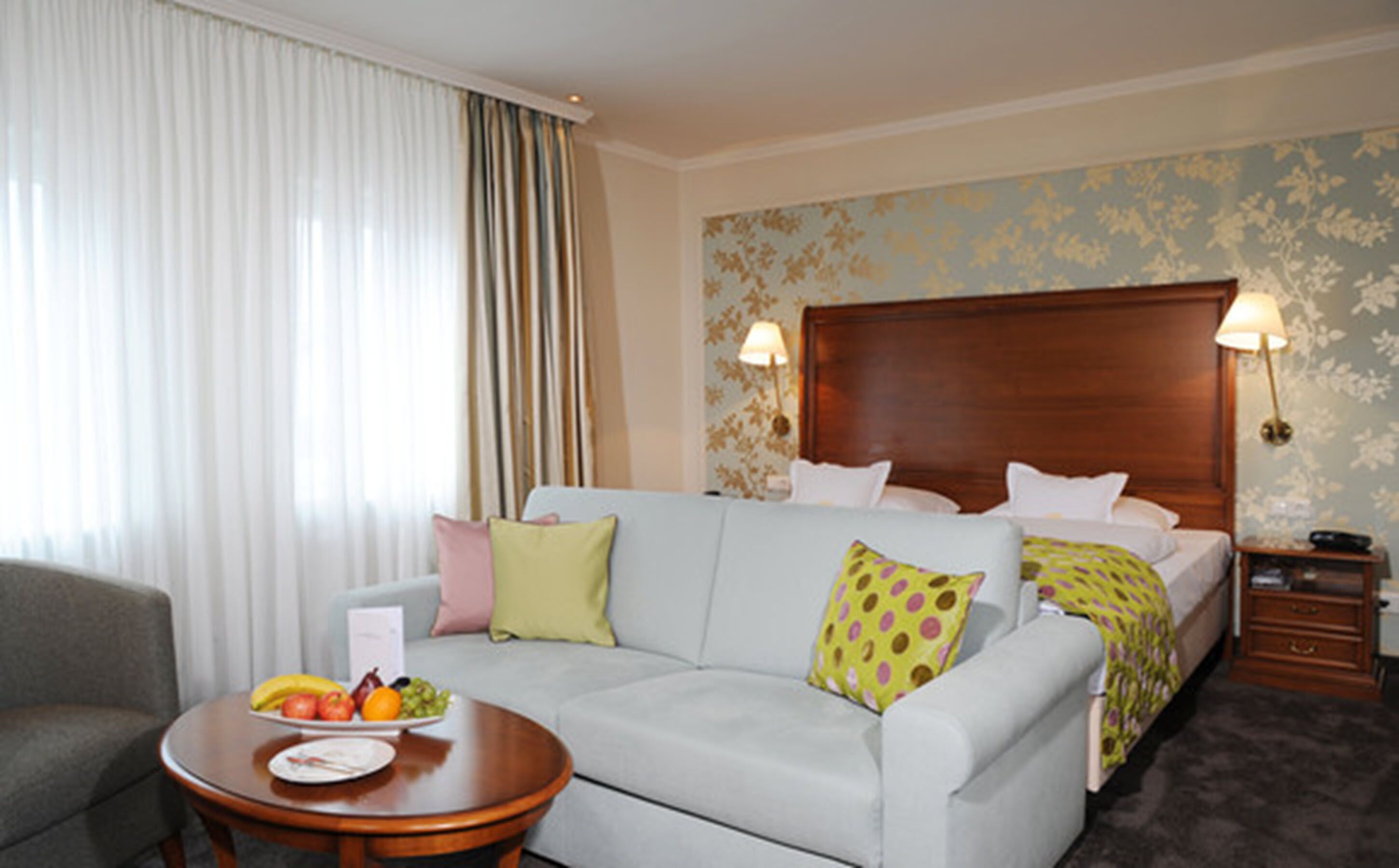 Habitación de hotel con cama, sofá, una mesita con fruta y una mesita de noche.