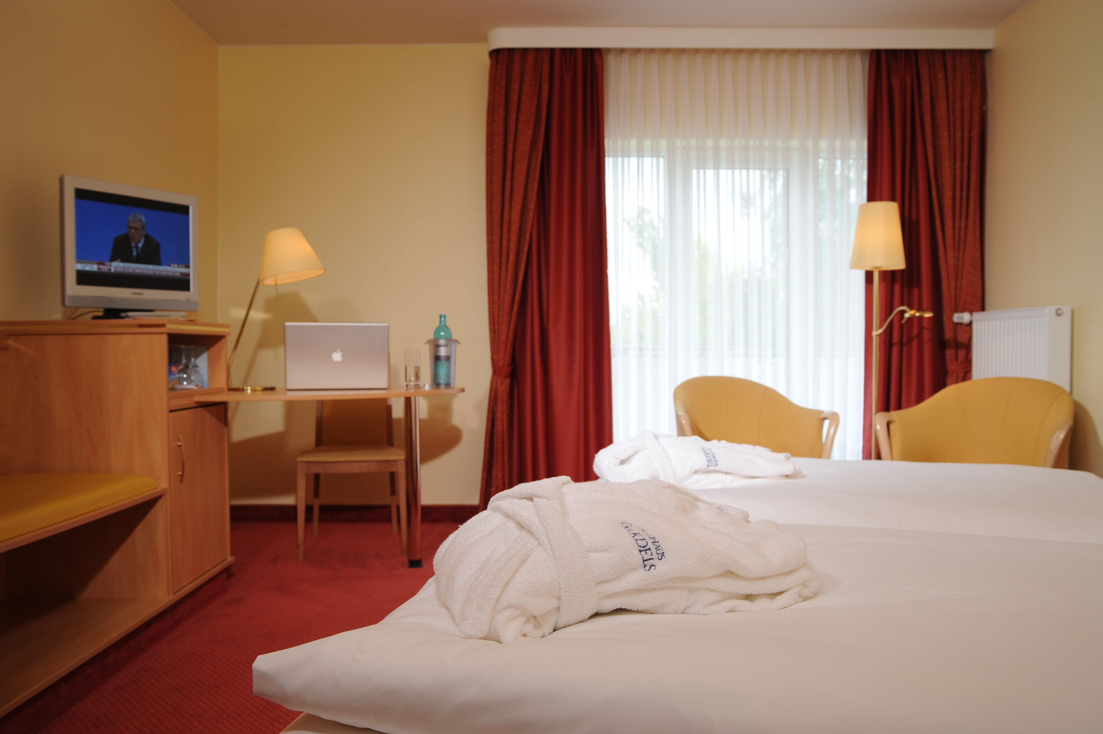 A hotel room with a bed, two chairs, a desk with a laptop, a television, and a radiator.
