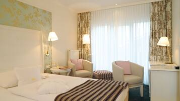 A hotel room with two beds, a sitting area with a chair and a small table, and a desk with a lamp.