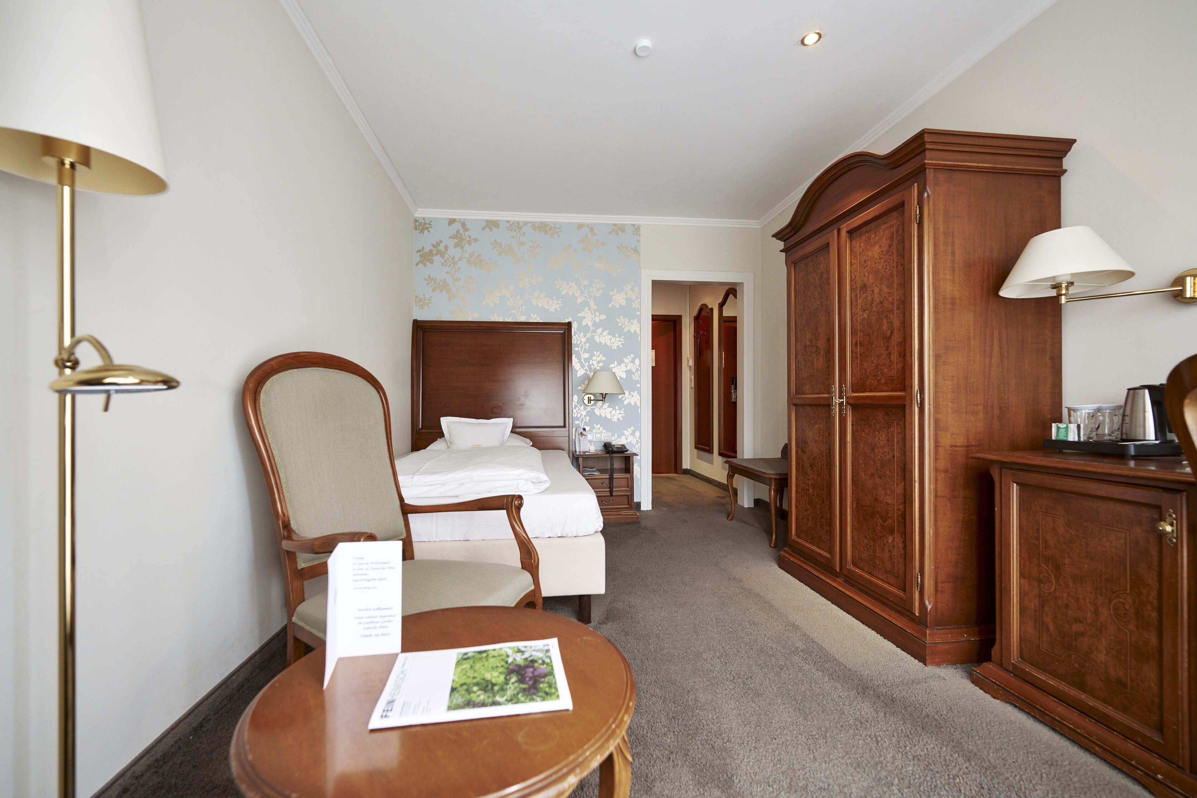 A hotel room with a bed, a wooden wardrobe, a desk with a lamp, and a chair.