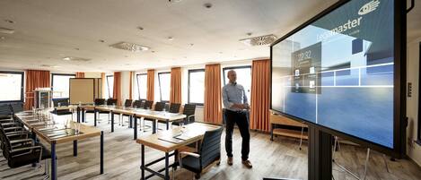 Un salón de conferencias con una pantalla grande que muestra Legamaster, una sala de reuniones con mesas, sillas y una pantalla para presentaciones.