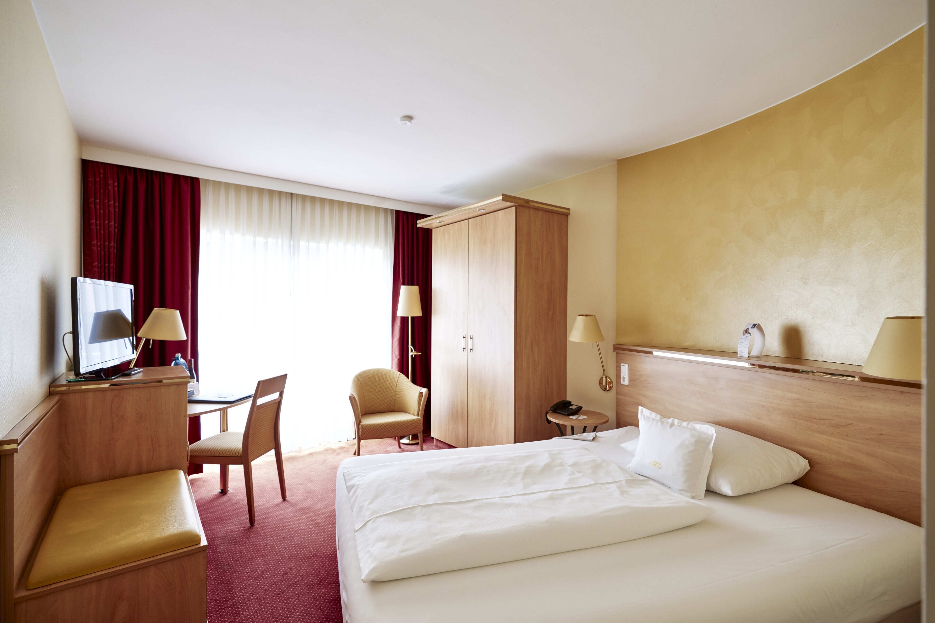 Ein Hotelzimmer mit Bett, Schreibtisch mit Computer, Stuhl, Kleiderschrank und Nachttisch mit Lampe.