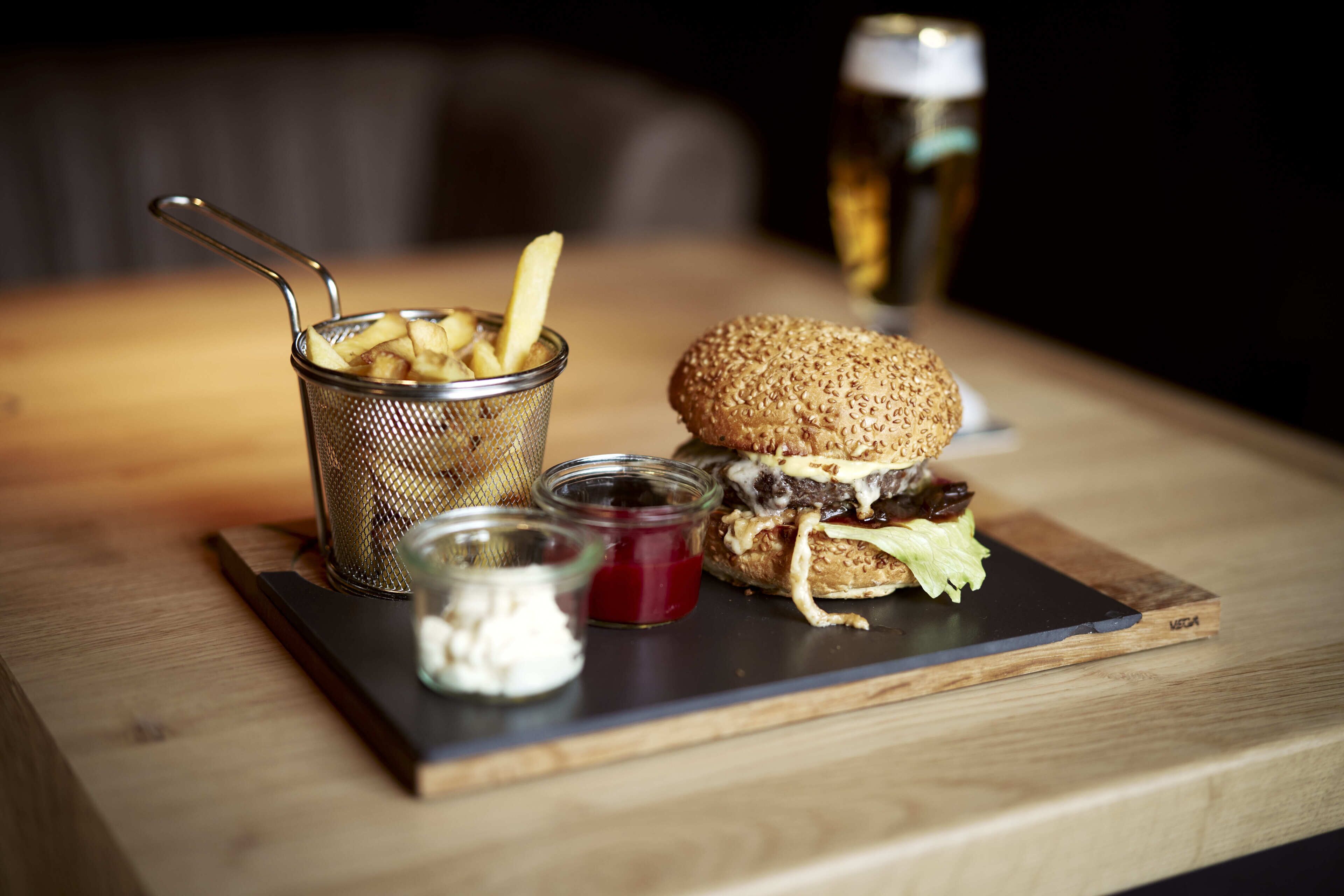 Hamburguesa con papas fritas y salsas sobre una tabla de madera, un vaso de cerveza y un colador metálico con papas fritas.