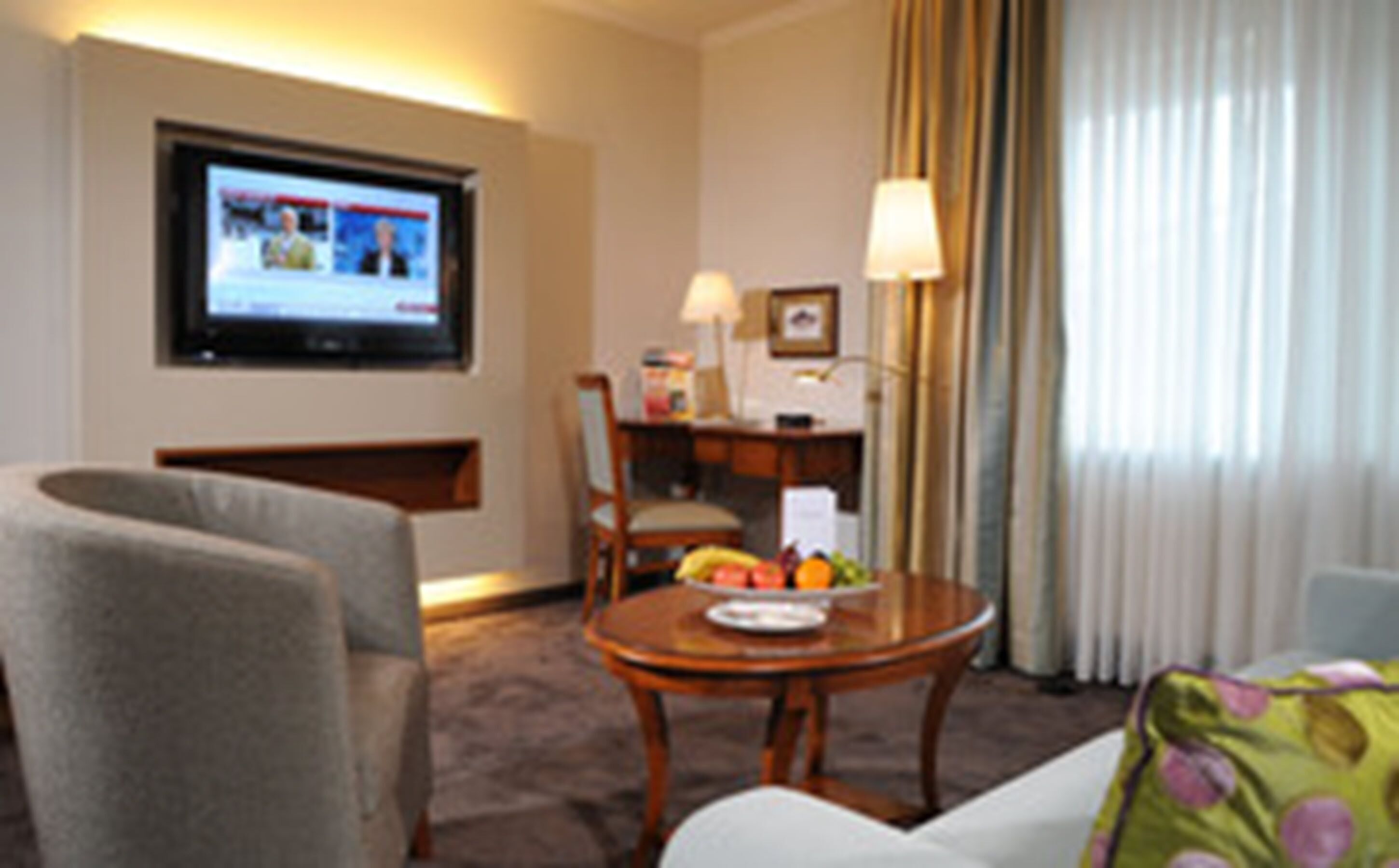 Een hotelkamer met een televisie, een bureau, een stoel, een eettafel met fruit en een bank.