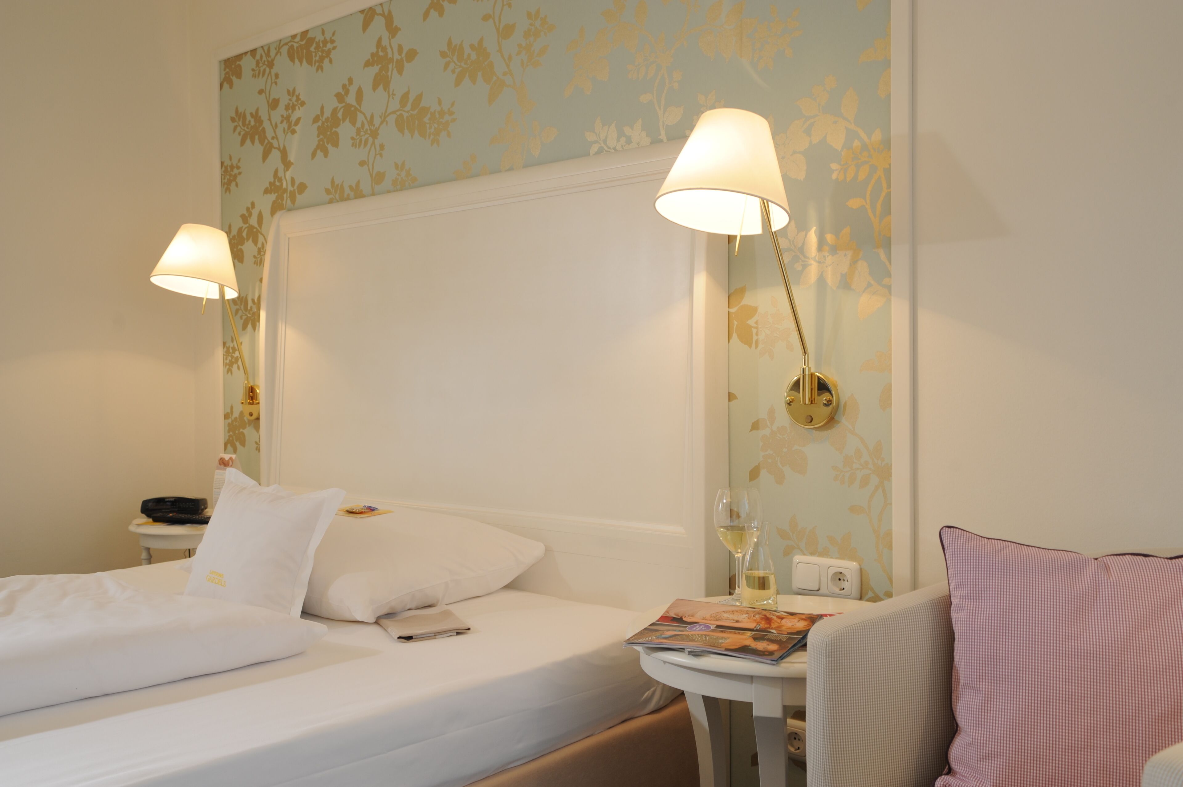 A hotel room with a bed, two bedside lamps, a nightstand, a magazine, and a wall with floral wallpaper.