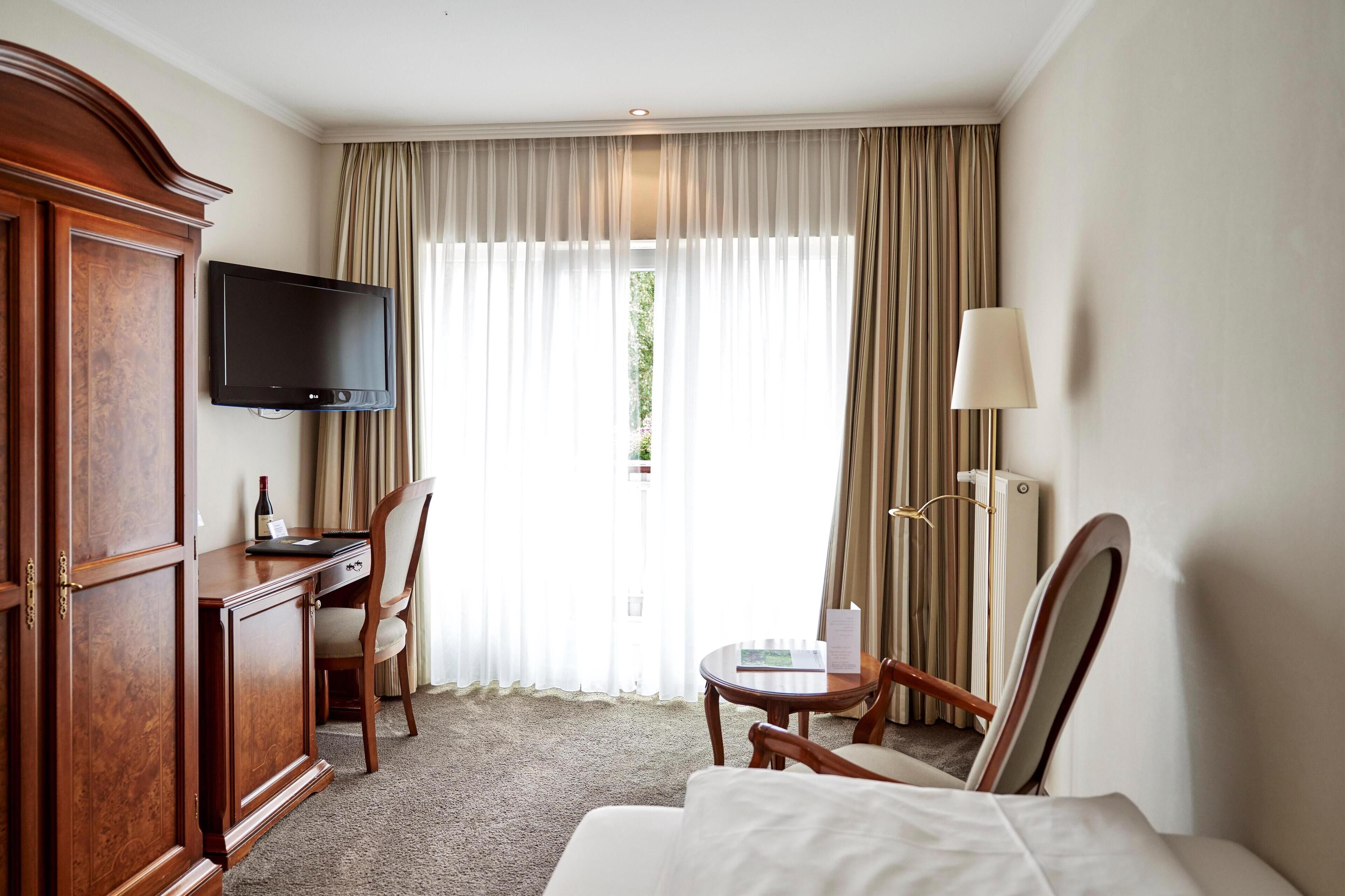 Habitación de hotel con un amplio armario de madera, un televisor de pantalla plana en la pared, un escritorio con silla, una mesita redonda con lámpara y una cama con edredón blanco.