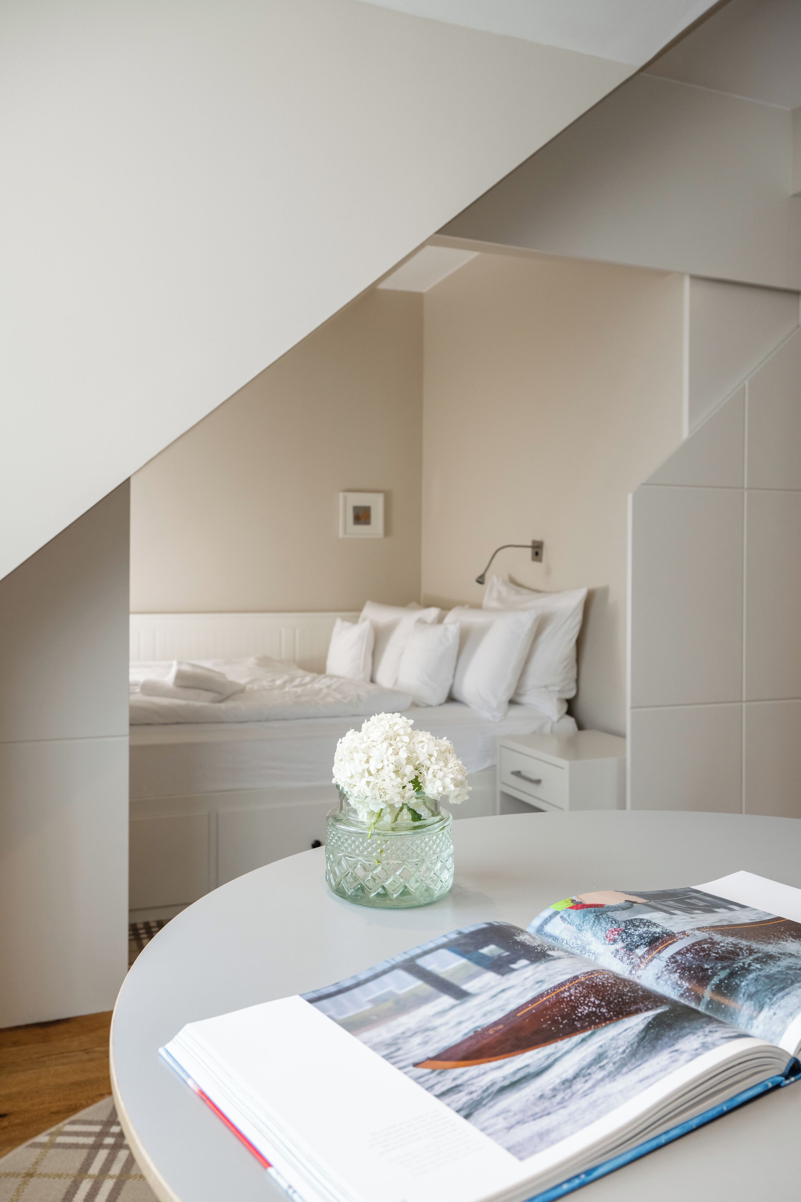 Ein modernes Schlafzimmer mit einem weißen Bett, einem Nachttisch und einer Vase mit weißen Blumen.