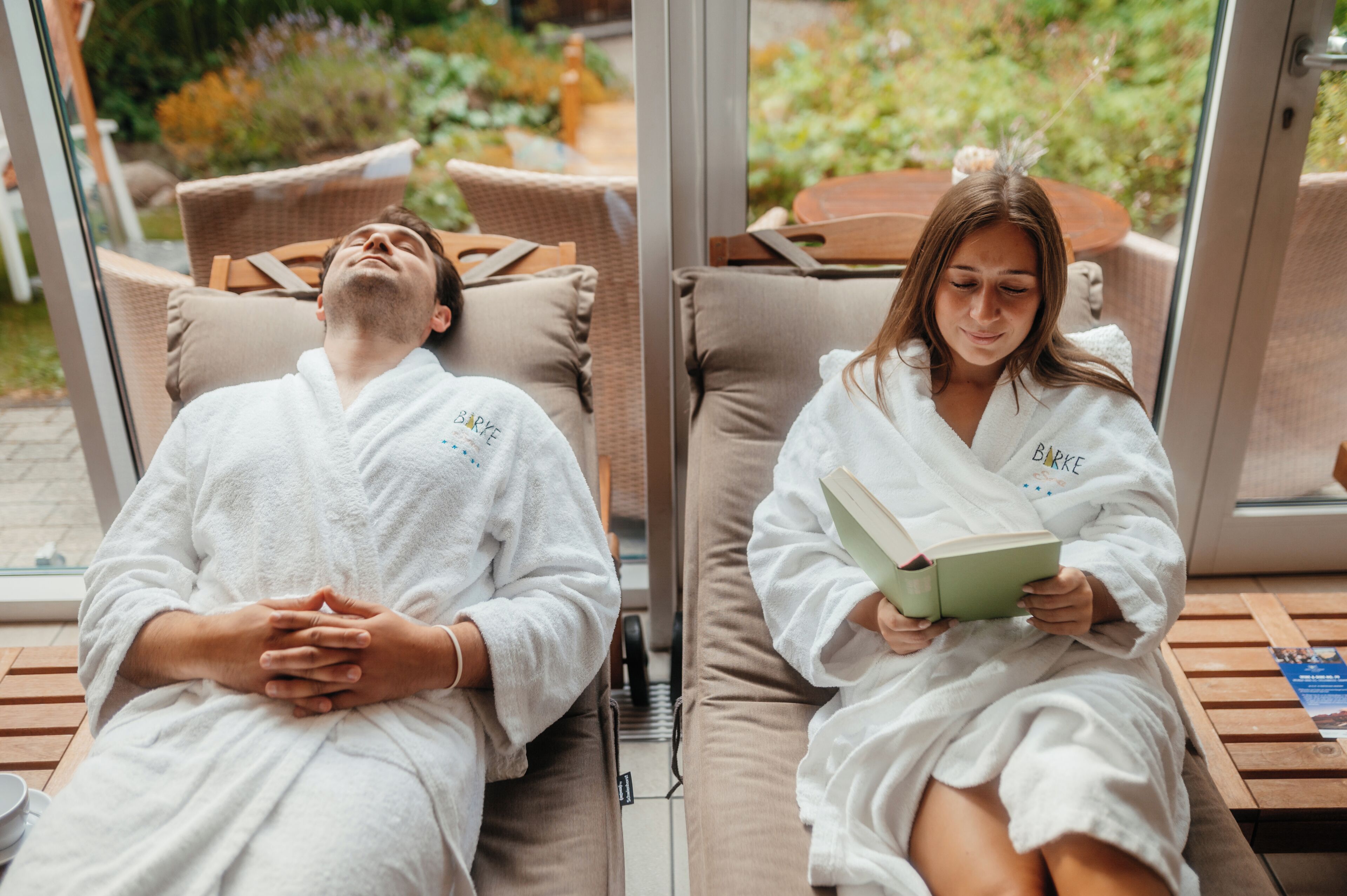 Two individuals in white bathrobes seated on outdoor lounge chairs with cushions, one reading a book and the other with eyes closed.