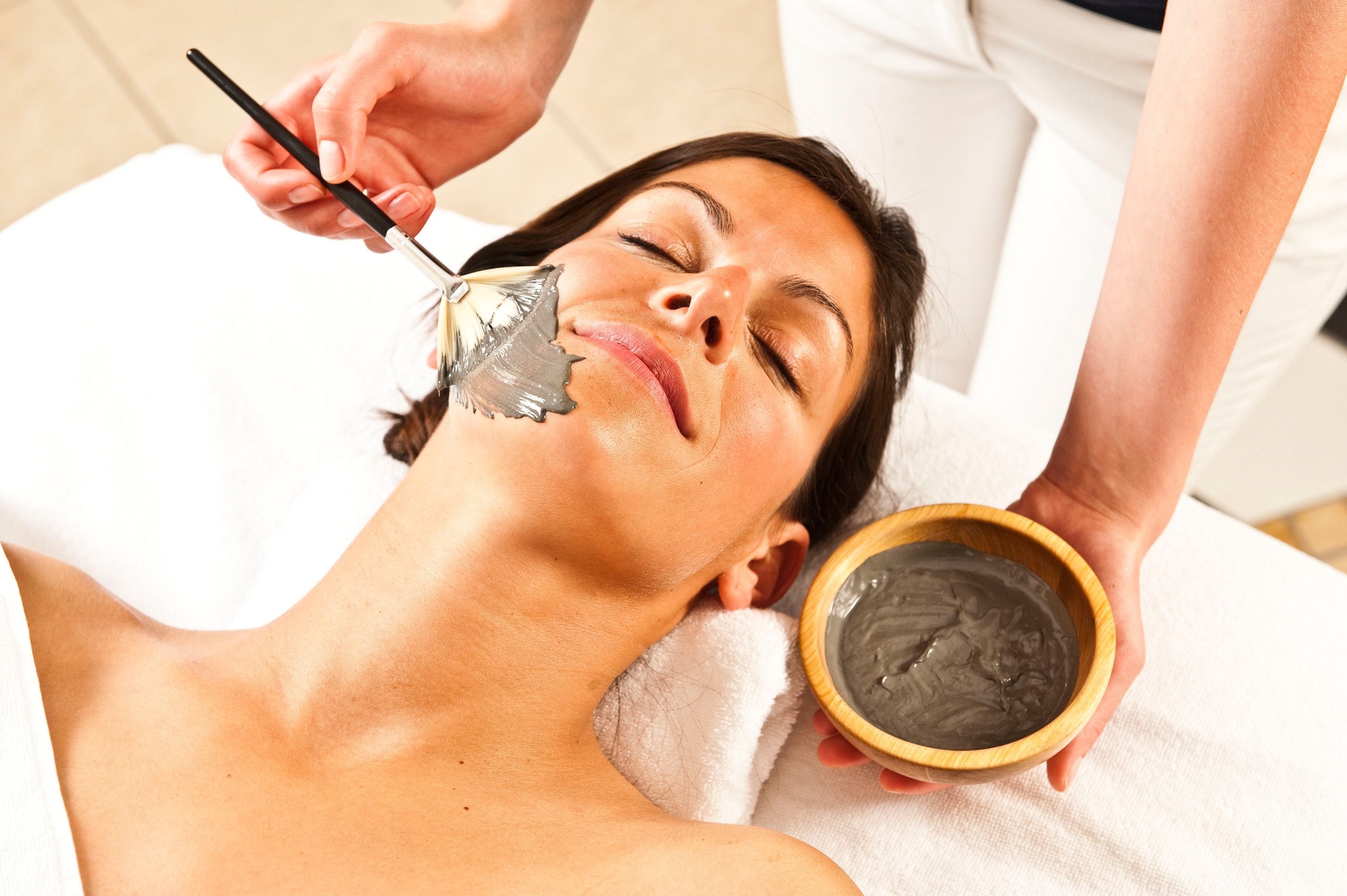 A person receiving a facial treatment with a brush and a bowl of product.