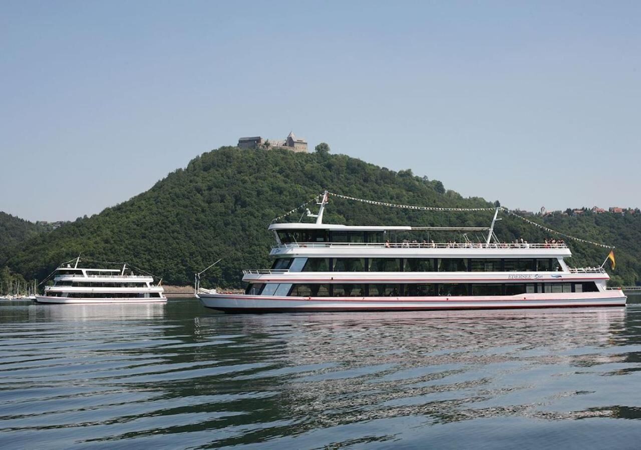 A large, multi-deck river cruise boat with a cabin structure, docked near a hill with a building on top.
