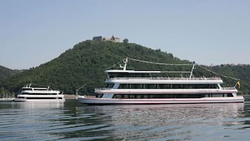A large, multi-deck river cruise boat with a cabin structure, docked near a hill with a building on top.