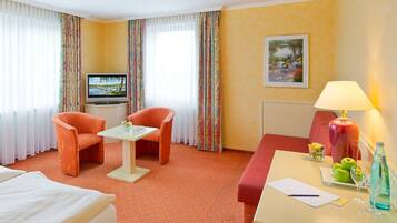 Une chambre d’hôtel avec un lit, deux fauteuils orange, une petite table, un téléviseur, un bureau avec une lampe, un tableau et une fenêtre avec des rideaux.