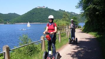 Dos personas en Segways por un sendero junto a un lago, con una colina y un edificio al fondo.