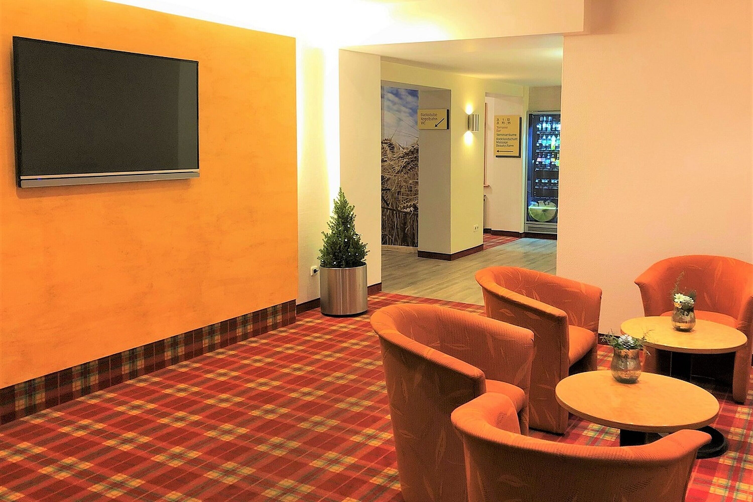 A lobby with a checkered floor, orange chairs, a small table with a vase, a potted plant, and a wall-mounted TV.