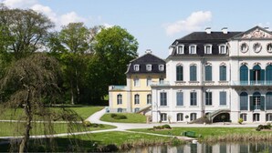 A large, multi-story building with a symmetrical facade, a central entrance with a portico, and a reflection in a body of water. - Waldhotel Schäferberg (Espenau)
