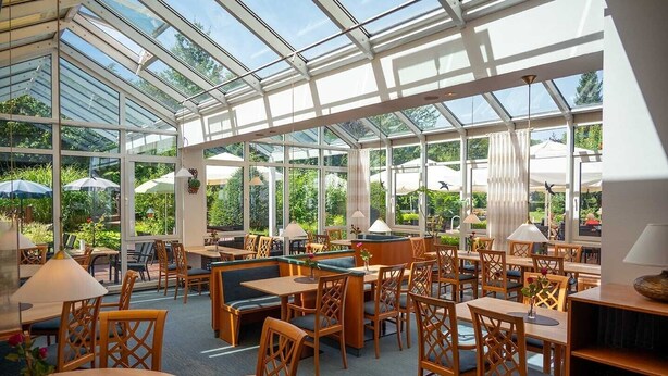 A conservatory with glass walls, wooden furniture, and outdoor views. - Waldhotel Schäferberg (Espenau)