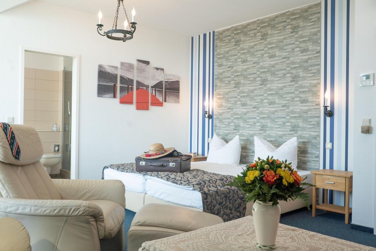 Ein Hotelzimmer mit einem Bett, einem Sessel, einem Koffer, einer Vase mit Blumen und einer Wand mit Streifenmuster.
