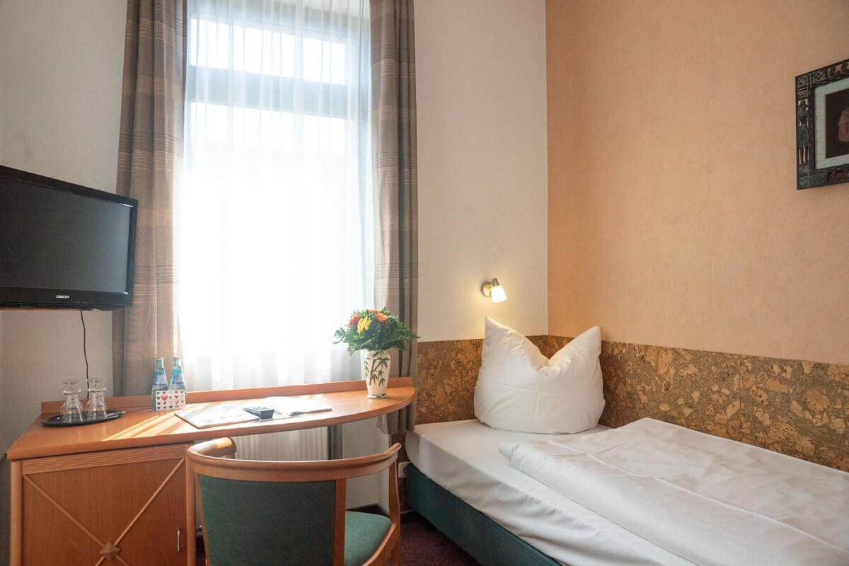 Ein Hotelzimmer mit einem Bett, einem Schreibtisch mit Stuhl, einem Fernseher, einem Fenster mit Vorhängen und einer Wandleuchte.