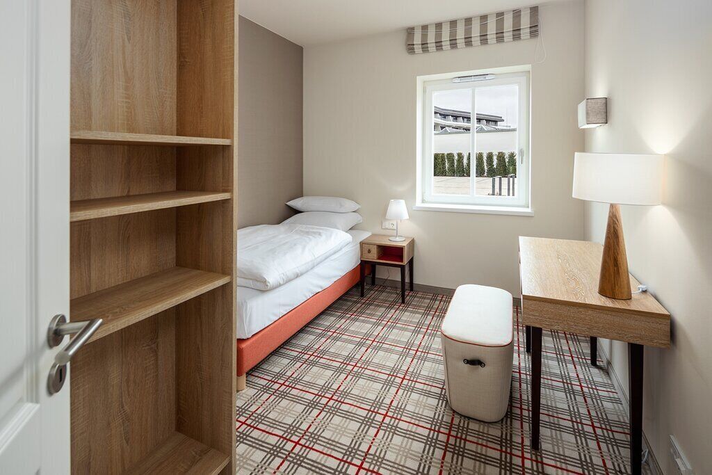 A hotel room with a bed, a bedside table with a lamp, a small stool, and a window with a view of buildings and trees.