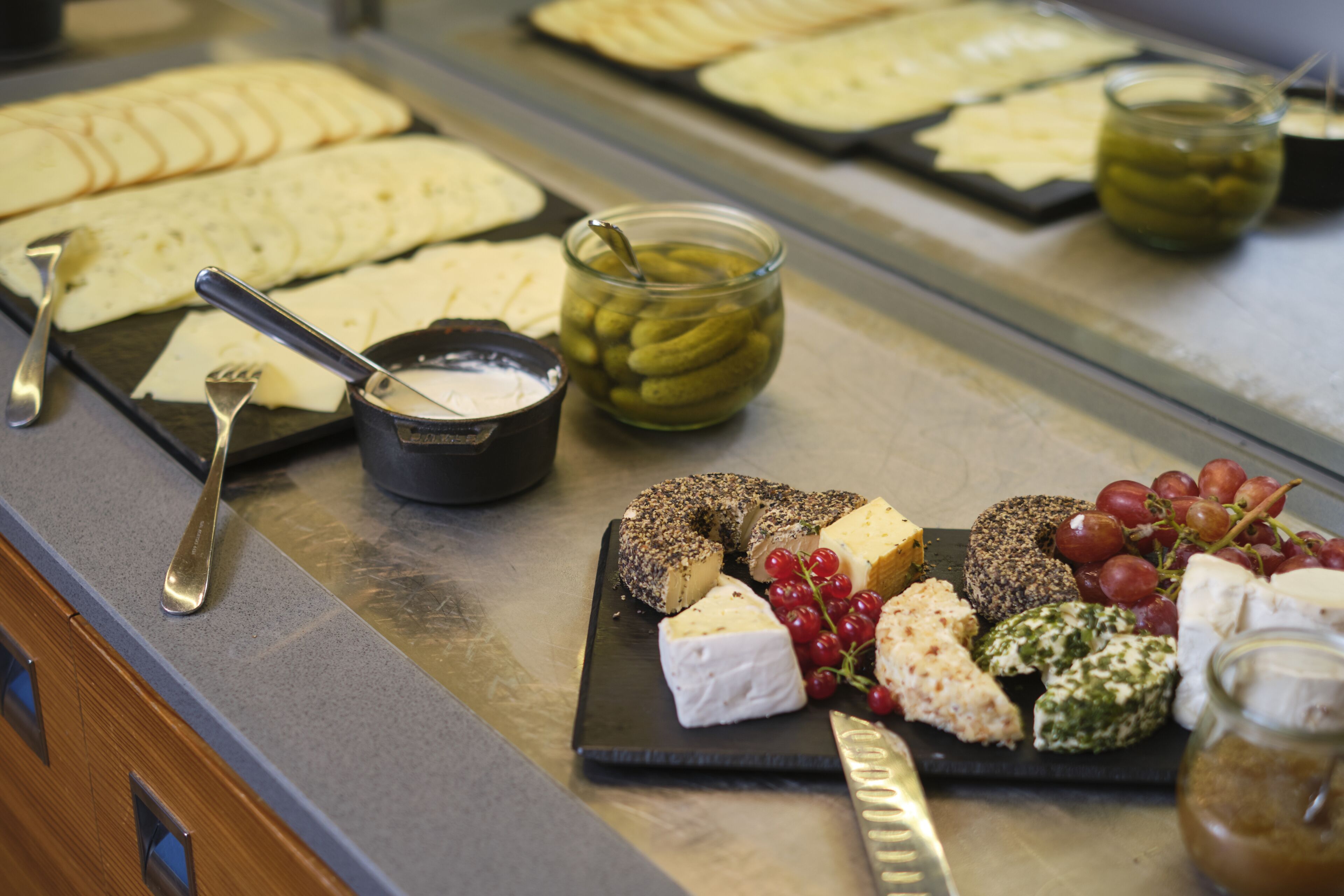 A cheese and cracker board with various cheeses, grapes, and a jar of pickles.