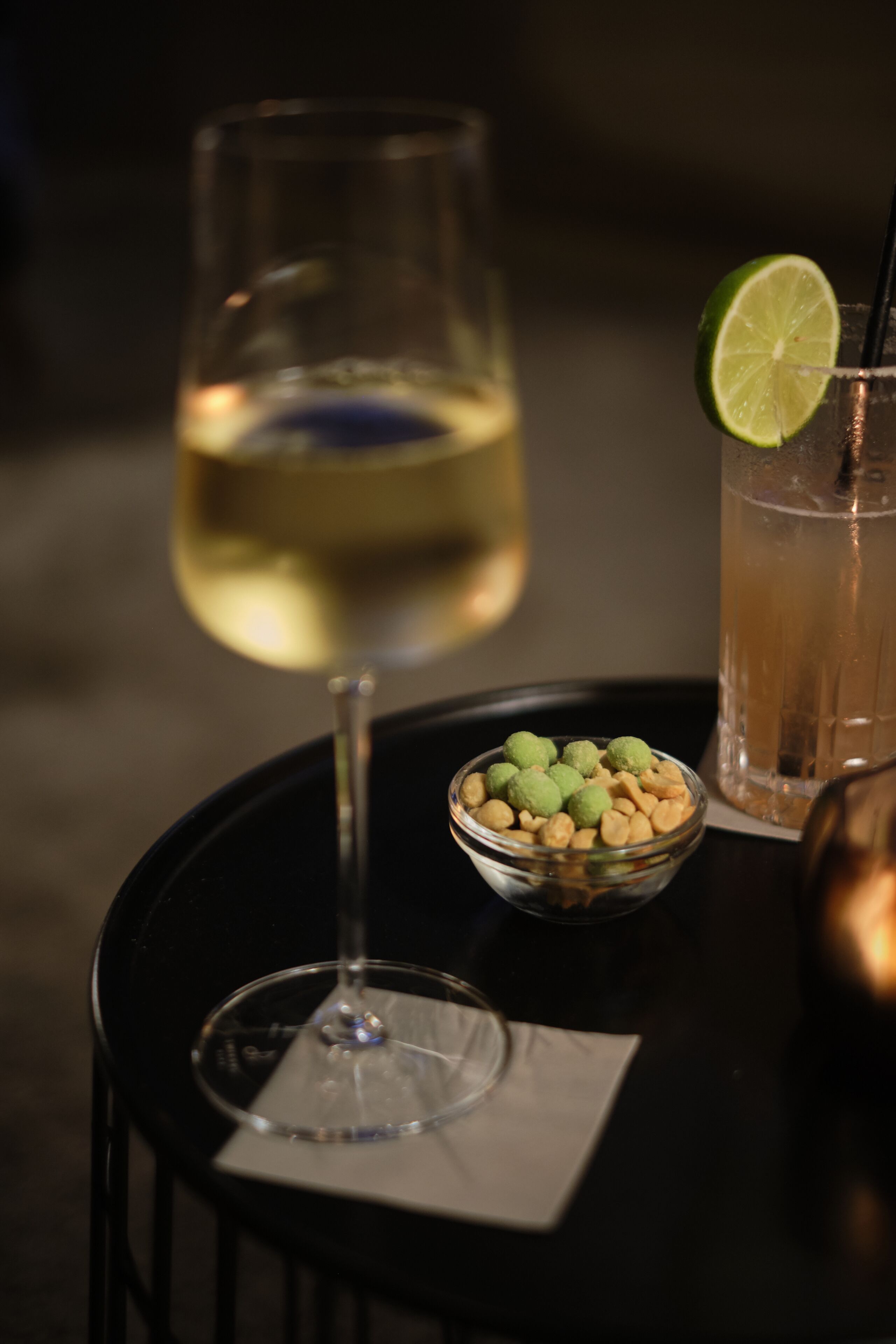 Ett glas vitt vin och en cocktail med en limeskiva på ett svart bord.