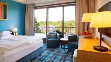 A hotel room with a bed, two armchairs, a desk with a lamp, a bowl of fruit, and a large window with a view of trees and buildings.