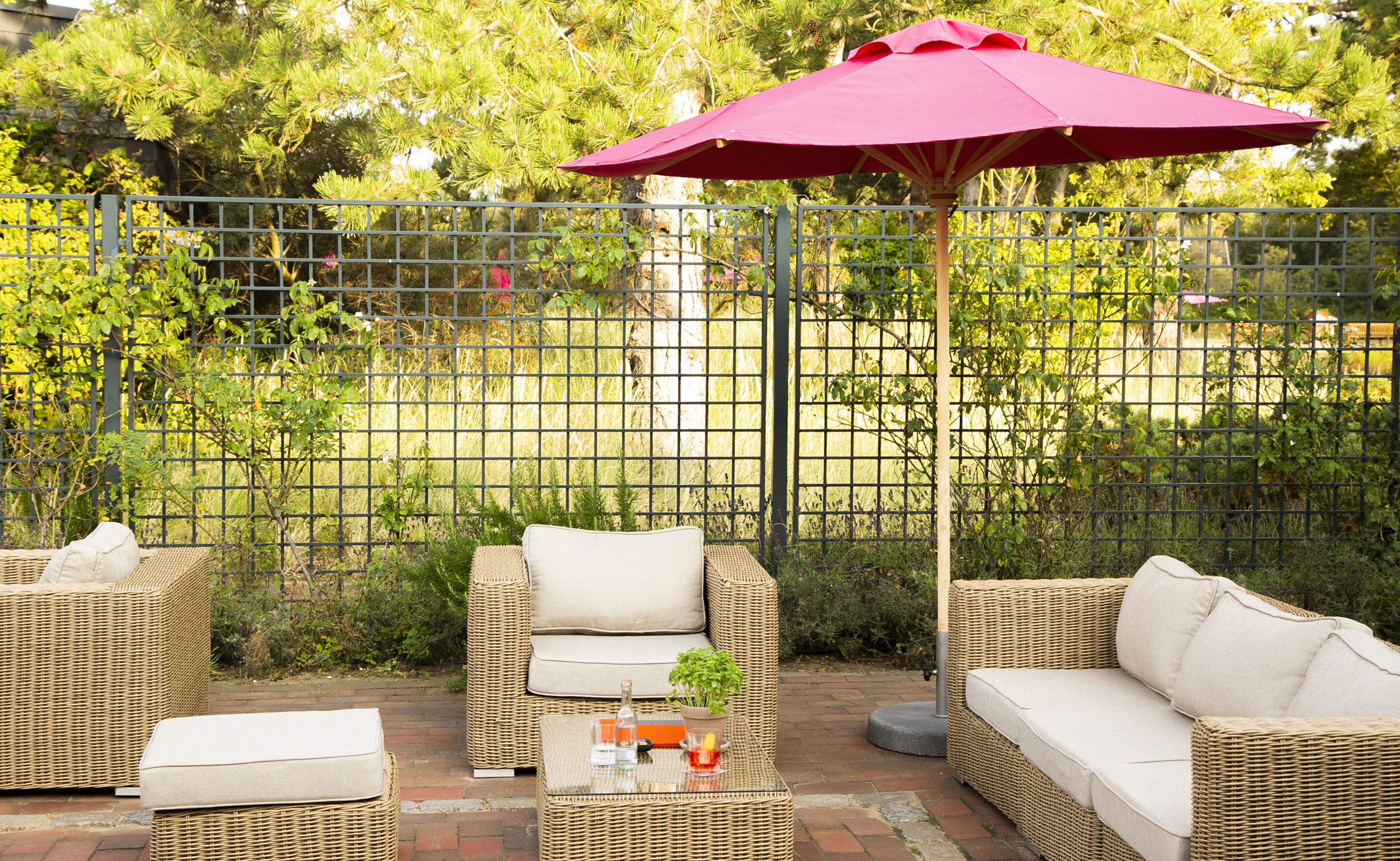 Un espace extérieur aménagé en terrasse, avec des meubles en osier, notamment des canapés et une table avec des boissons, et un parasol rouge pour faire de l’ombre.