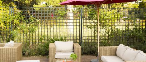 Un espace extérieur aménagé en terrasse, avec des meubles en osier, notamment des canapés et une table avec des boissons, et un parasol rouge pour faire de l’ombre.