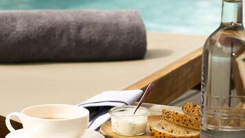 A wooden table with a cup, a small bowl of yogurt, a plate with bread, and a bottle of water by a poolside.