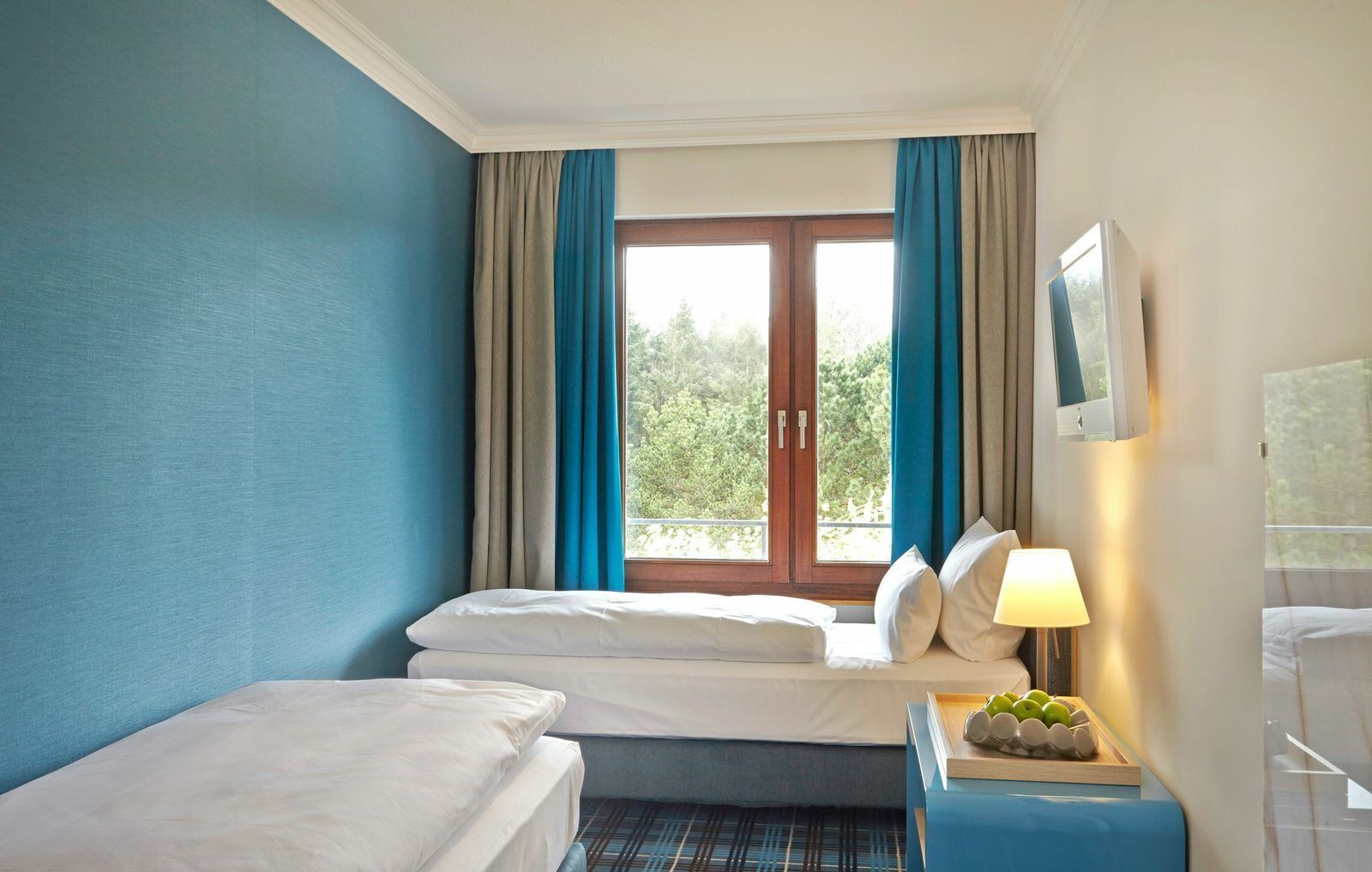 Una camera d'albergo con due letti, una finestra con tende e un comodino con una lampada, e vista su del verde all'esterno.