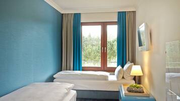 Ein Hotelzimmer mit zwei Betten, einem Fenster mit Vorhängen, einem Nachttisch mit Lampe und Blick auf Grünflächen.