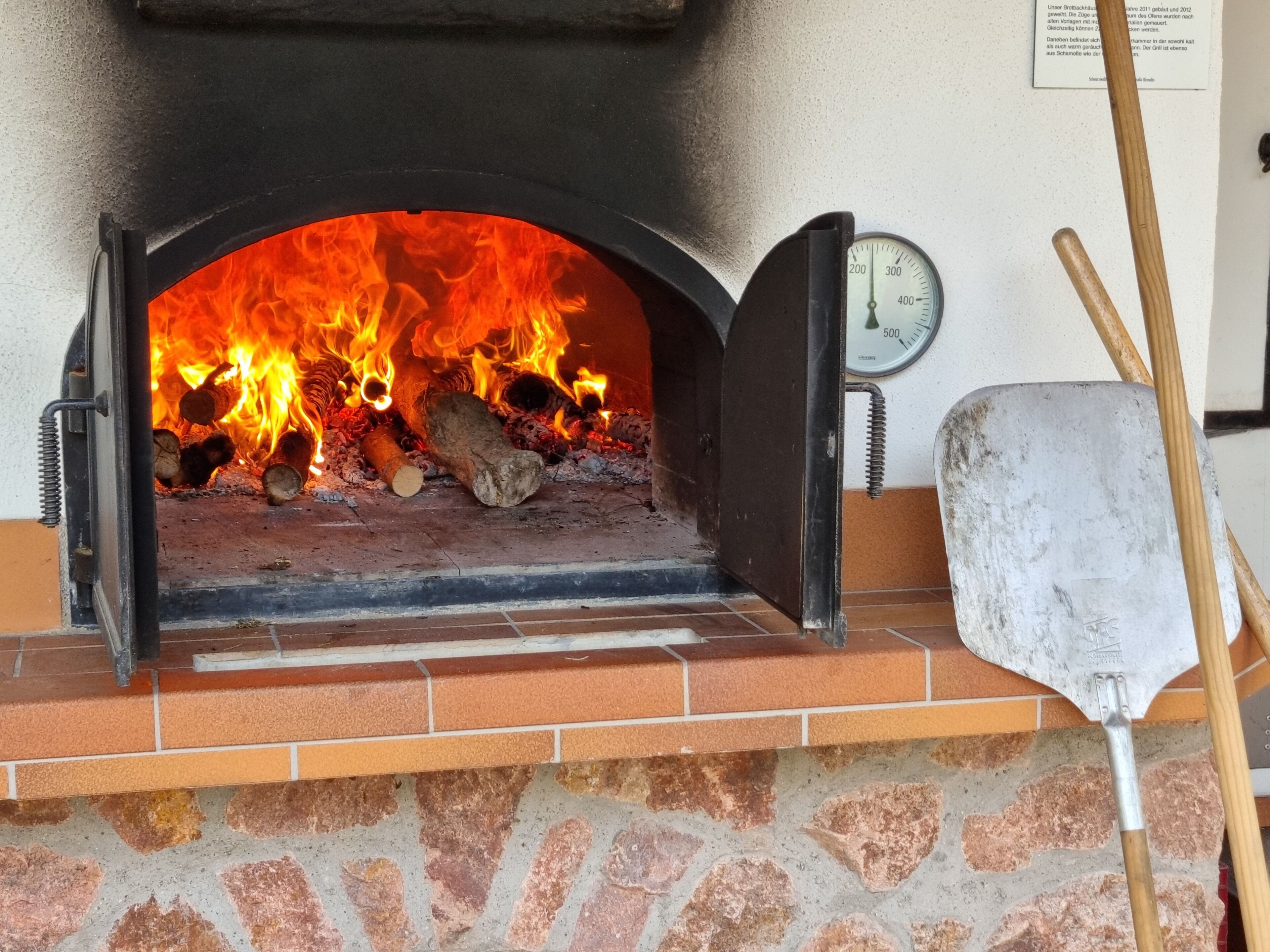 A brick oven with a fire burning inside, a thermometer, and a shovel.
