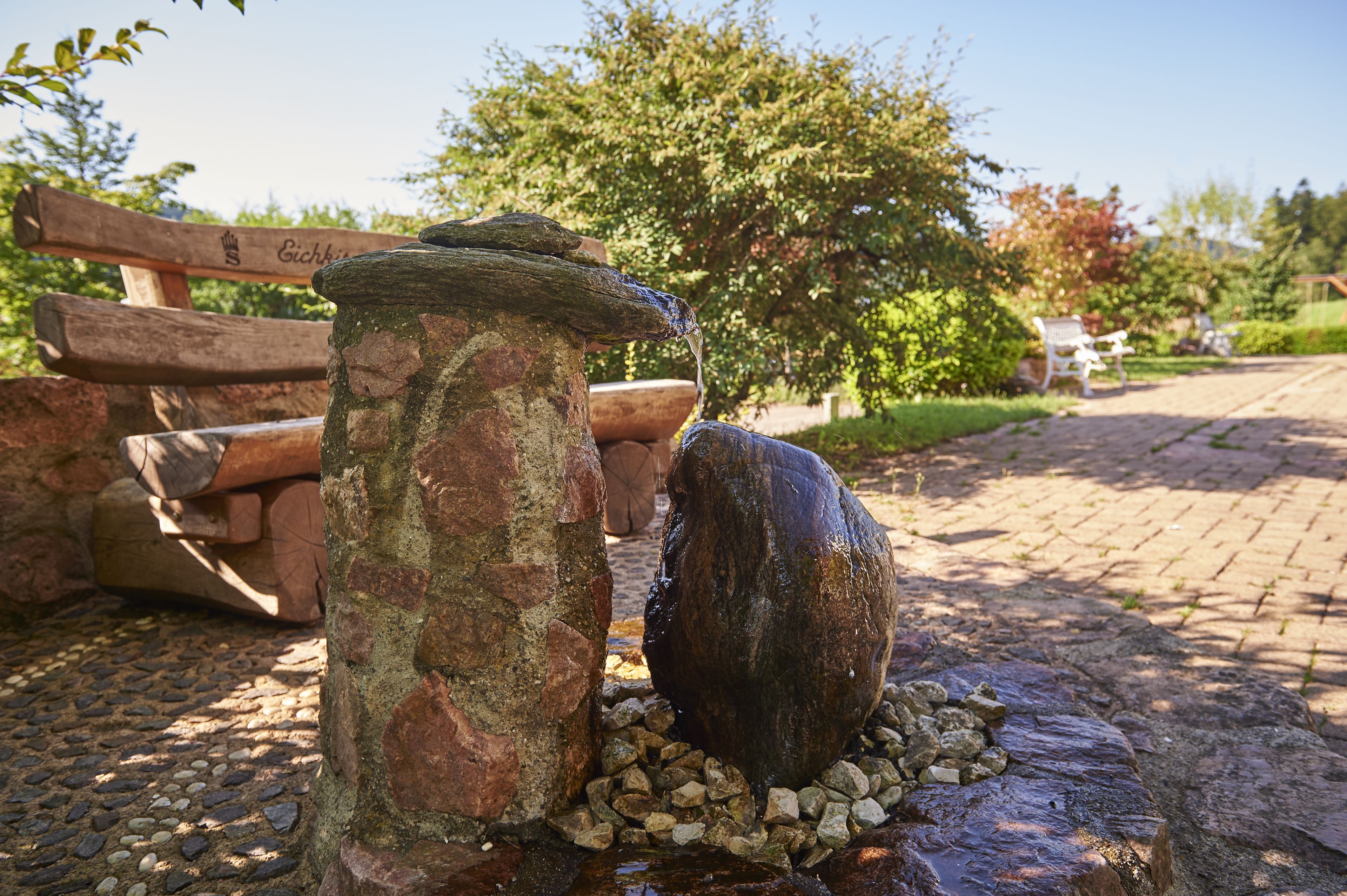 Een stenen fontein met stromend water, omringd door rotsen en een houten bank.