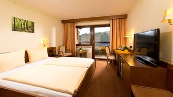 A hotel room with a large bed, a desk with a television, and a view of a forested area through the window.