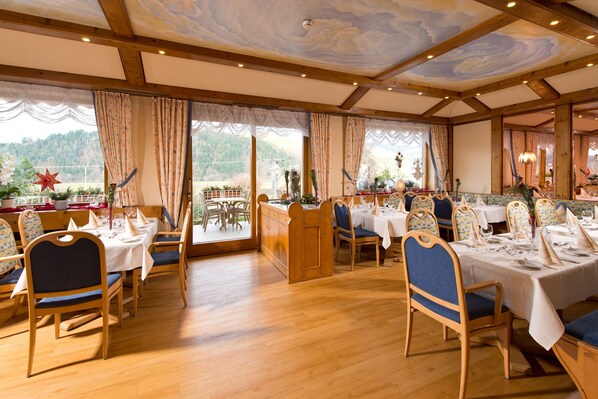 A restaurant with wooden tables and chairs, large windows with curtains, and a view of the outdoors.