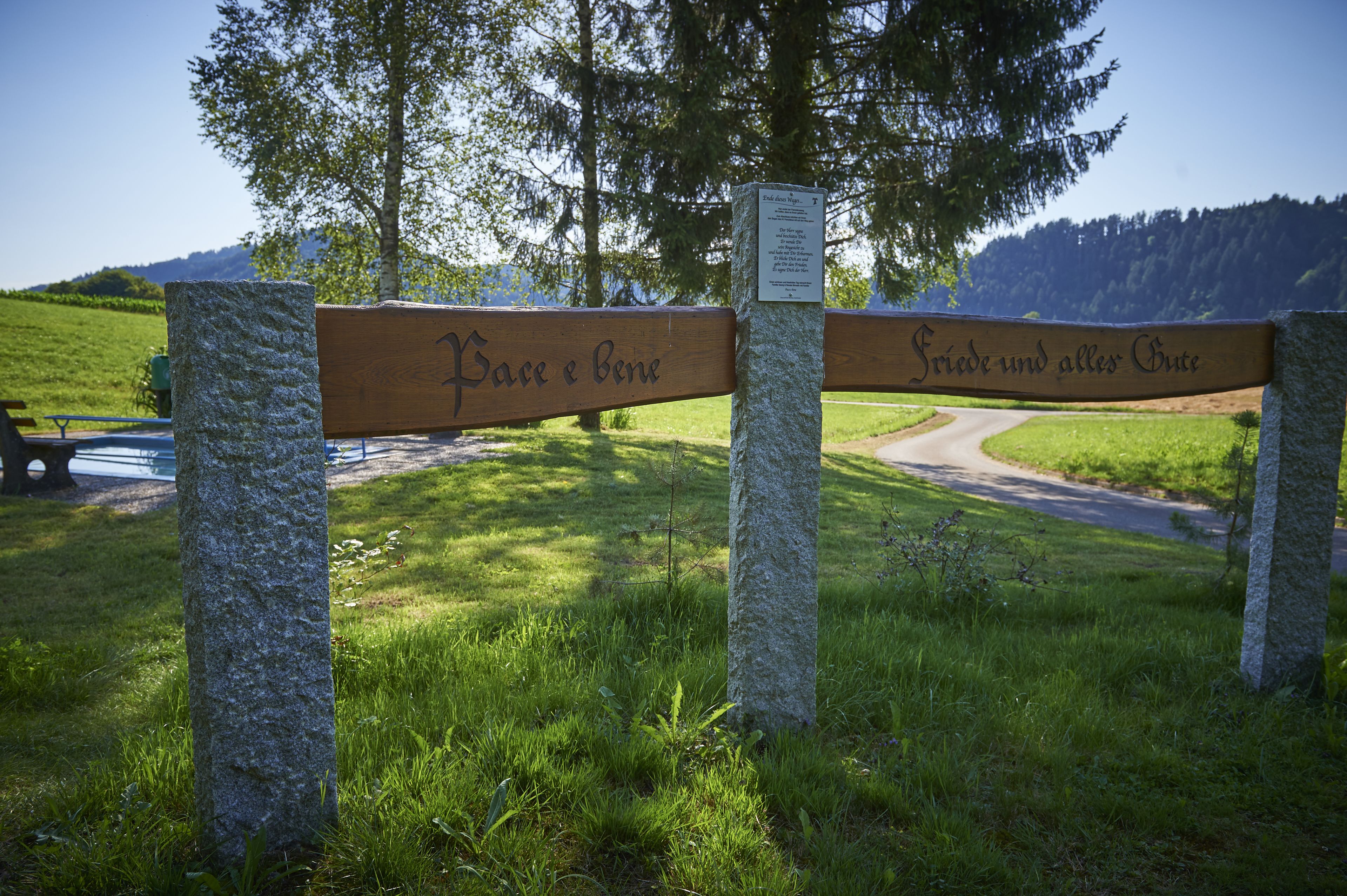 A wooden sign with inscriptions, a stone pillar, and a grassy area with a path and trees in the background.