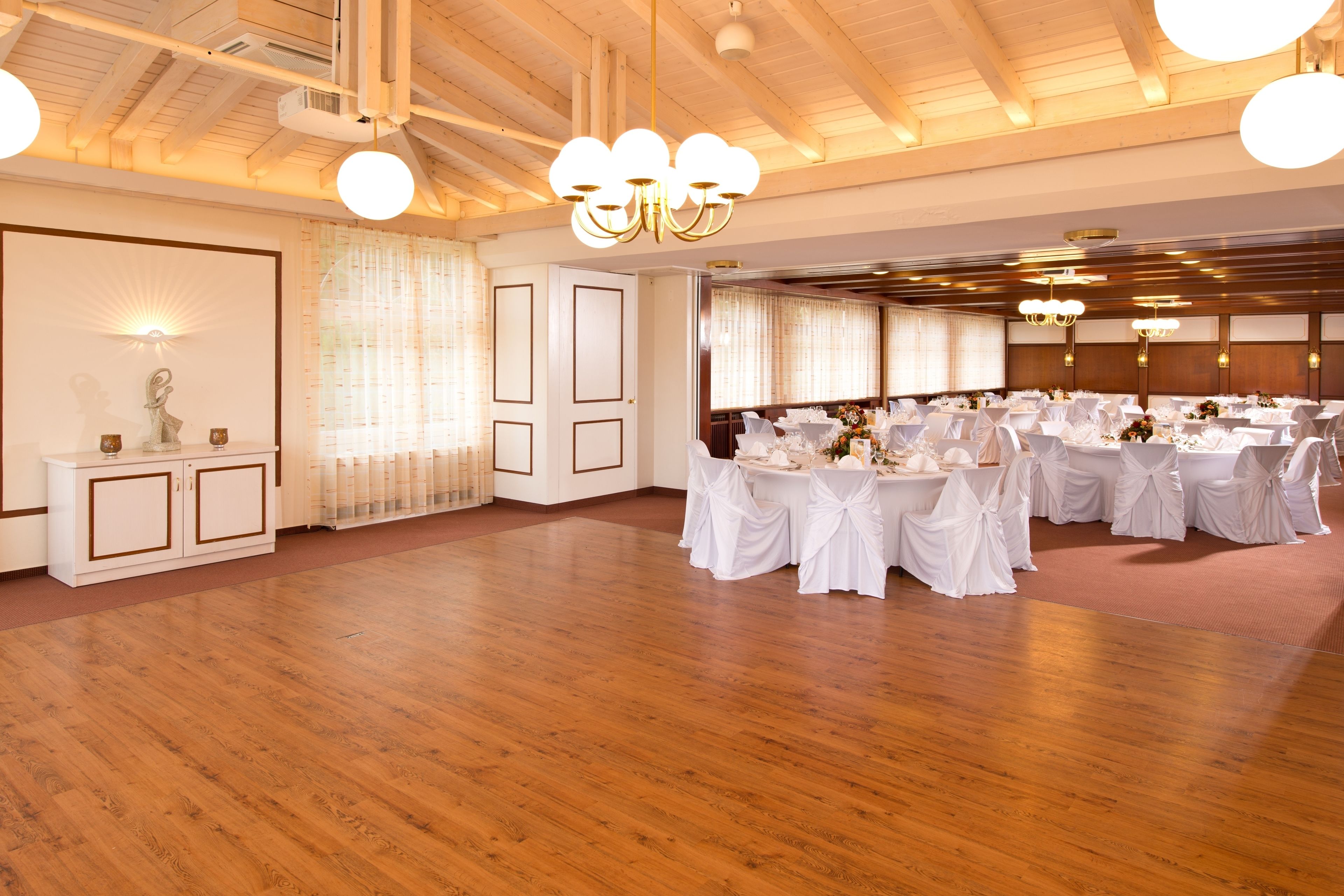 Une grande pièce dotée de parquets en bois, de tables blanches recouvertes de nappes blanches et de lustres.