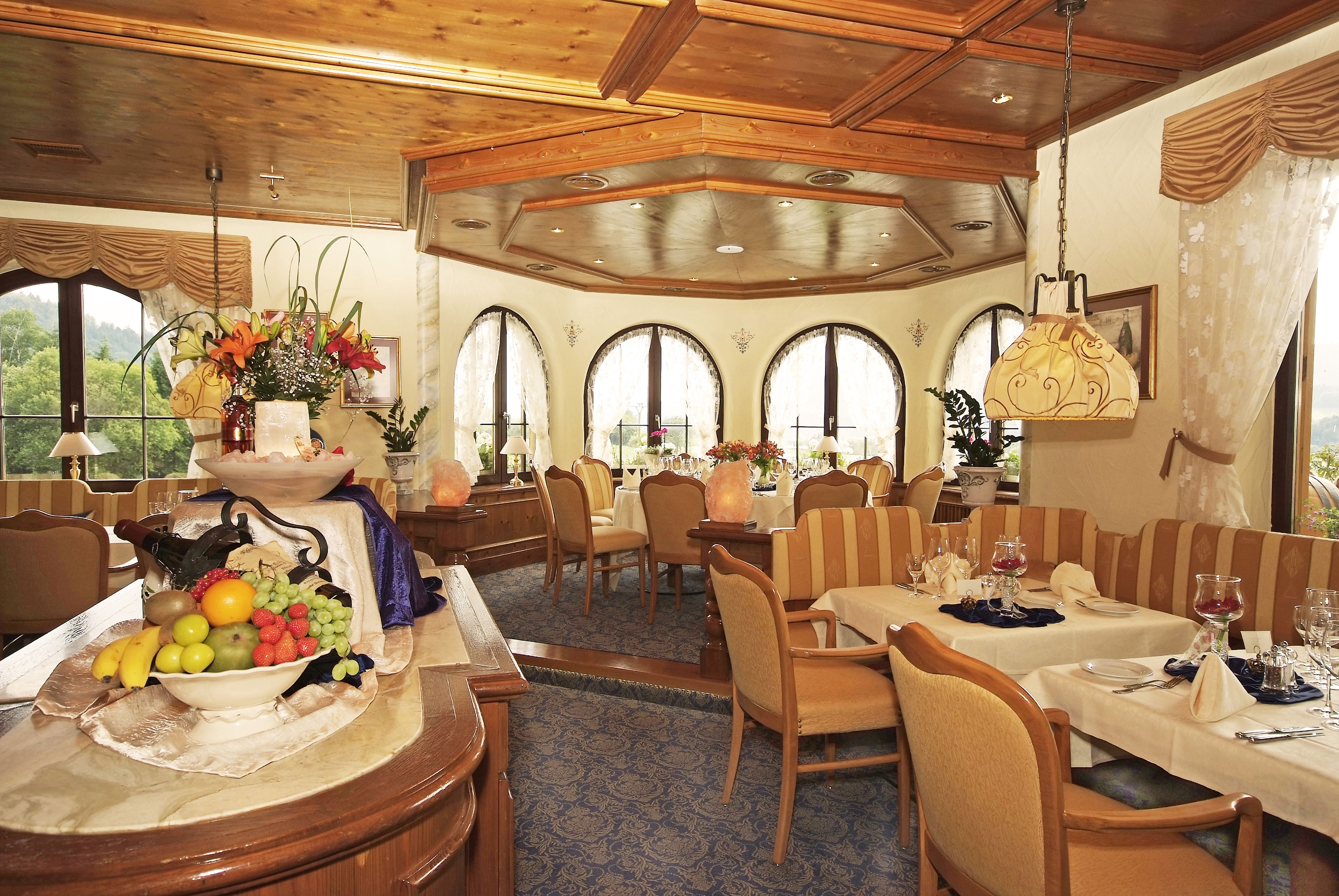 A dining area with wooden furniture, a fruit and vegetable centerpiece, and large arched windows.