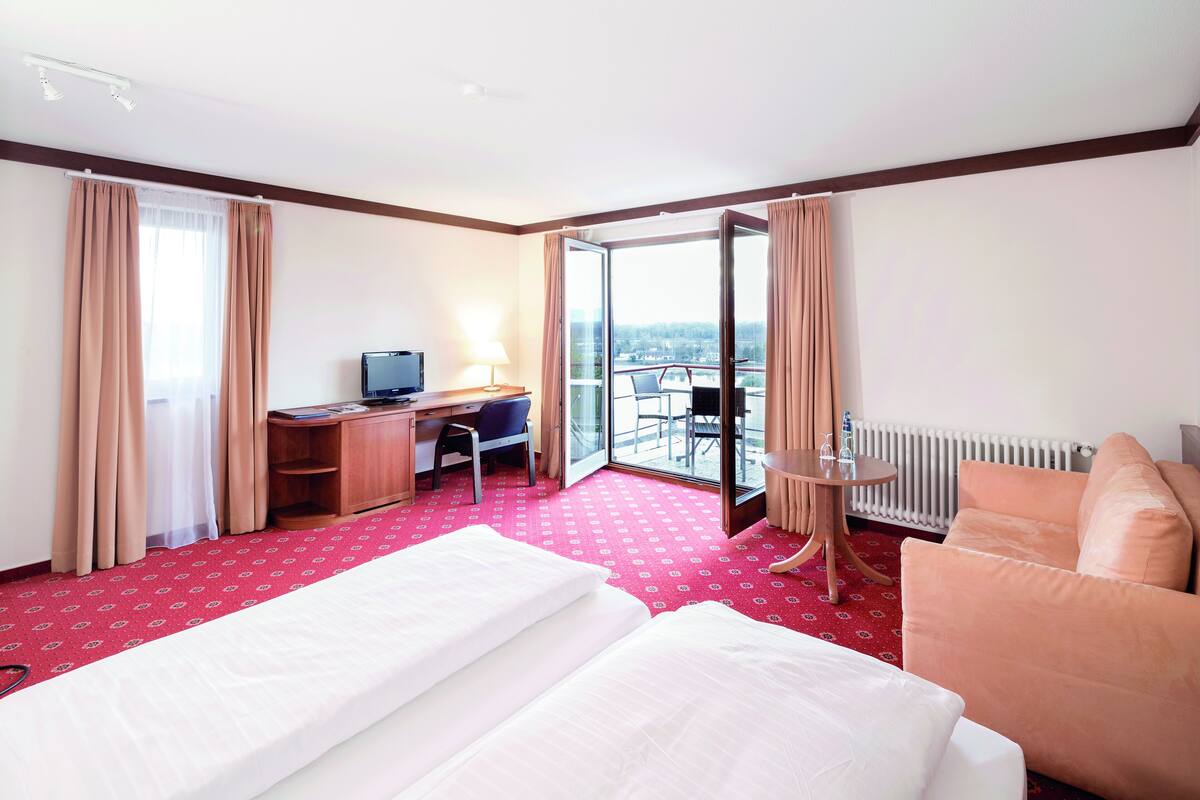 A hotel room with two beds, a desk with a computer, a chair, a small table, and a view of the outdoors.