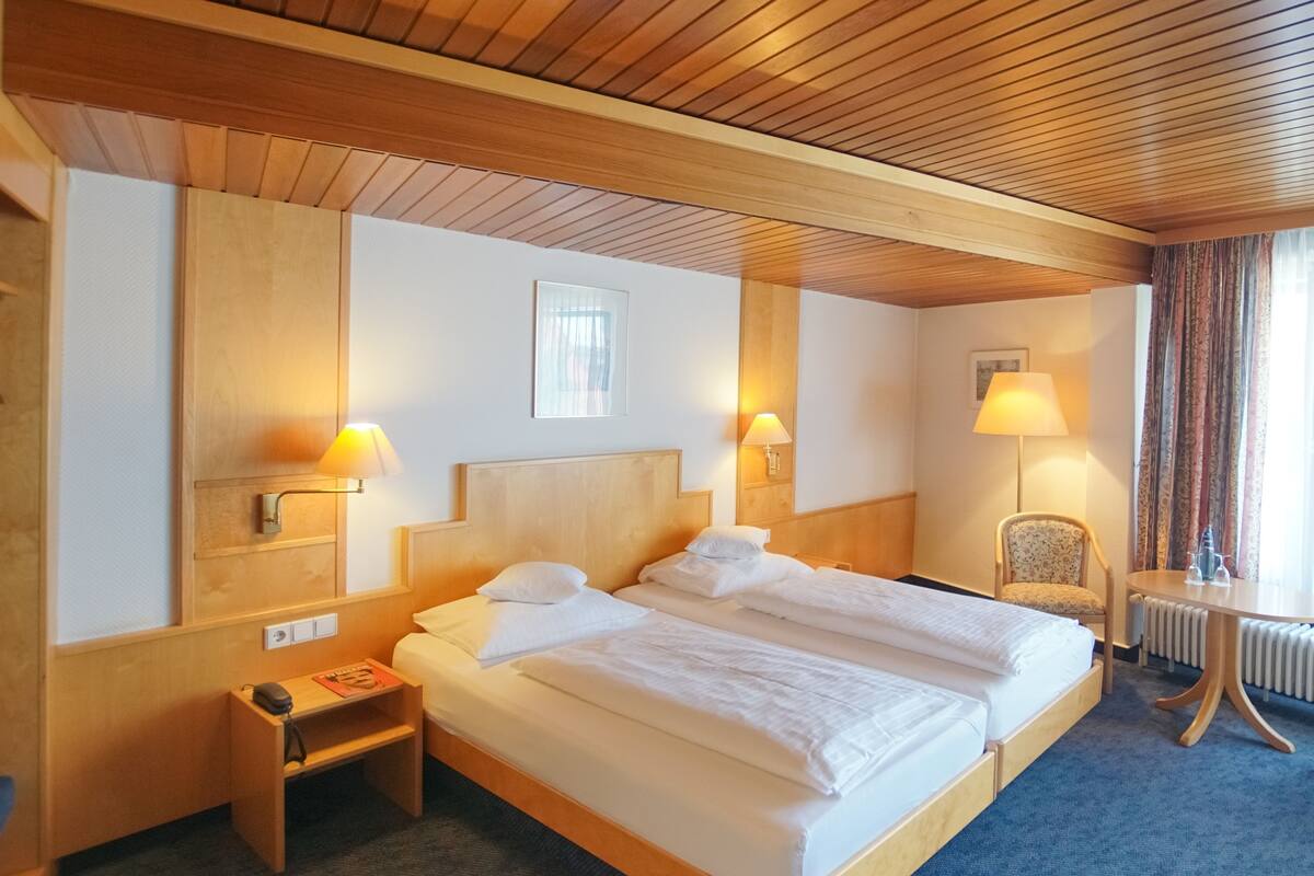 A hotel room with a wooden ceiling, a double bed with white linens, two bedside lamps, a chair, and a small table with a lamp.