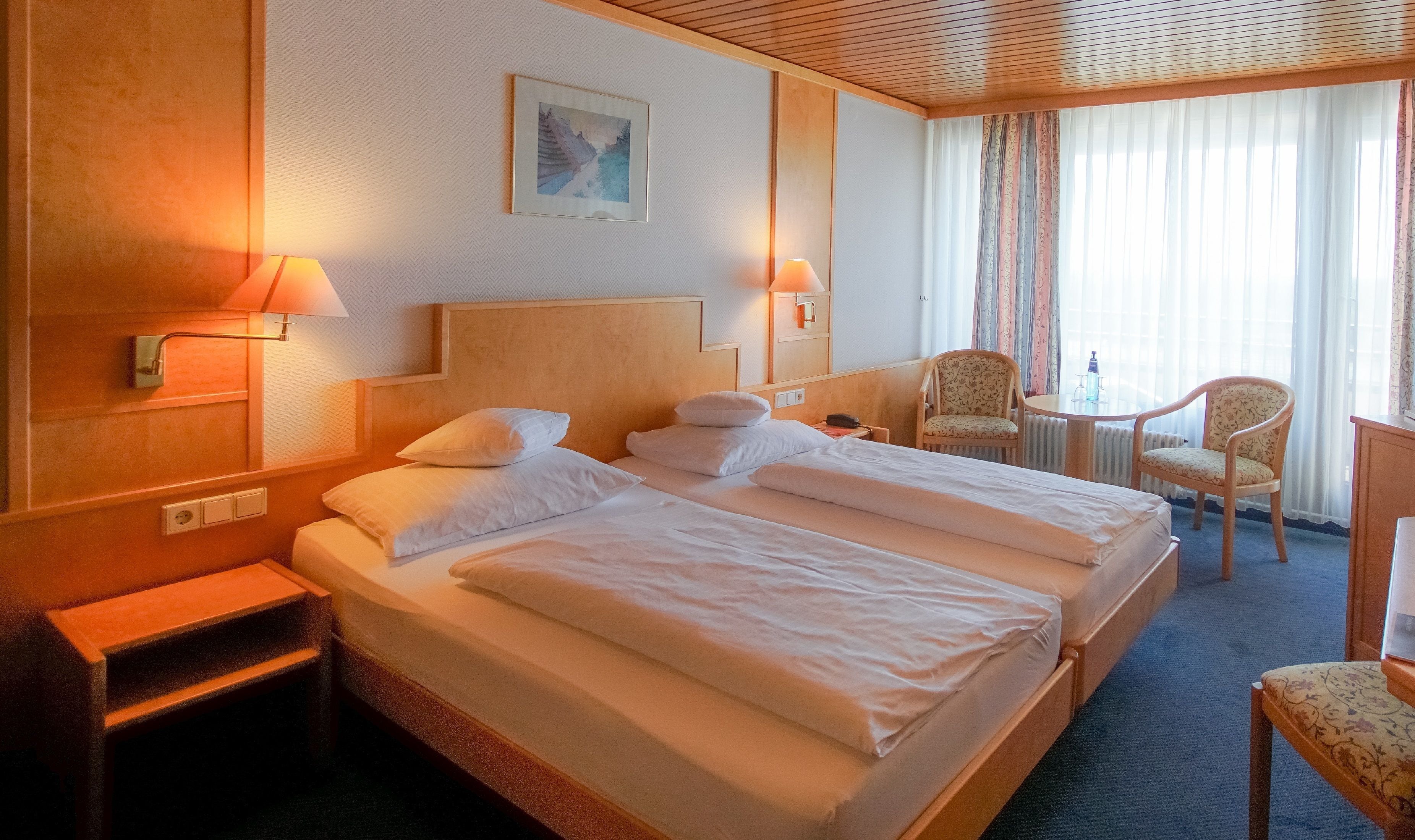 A hotel room with a large bed, two bedside tables, a chair, a small table, and a window with curtains.