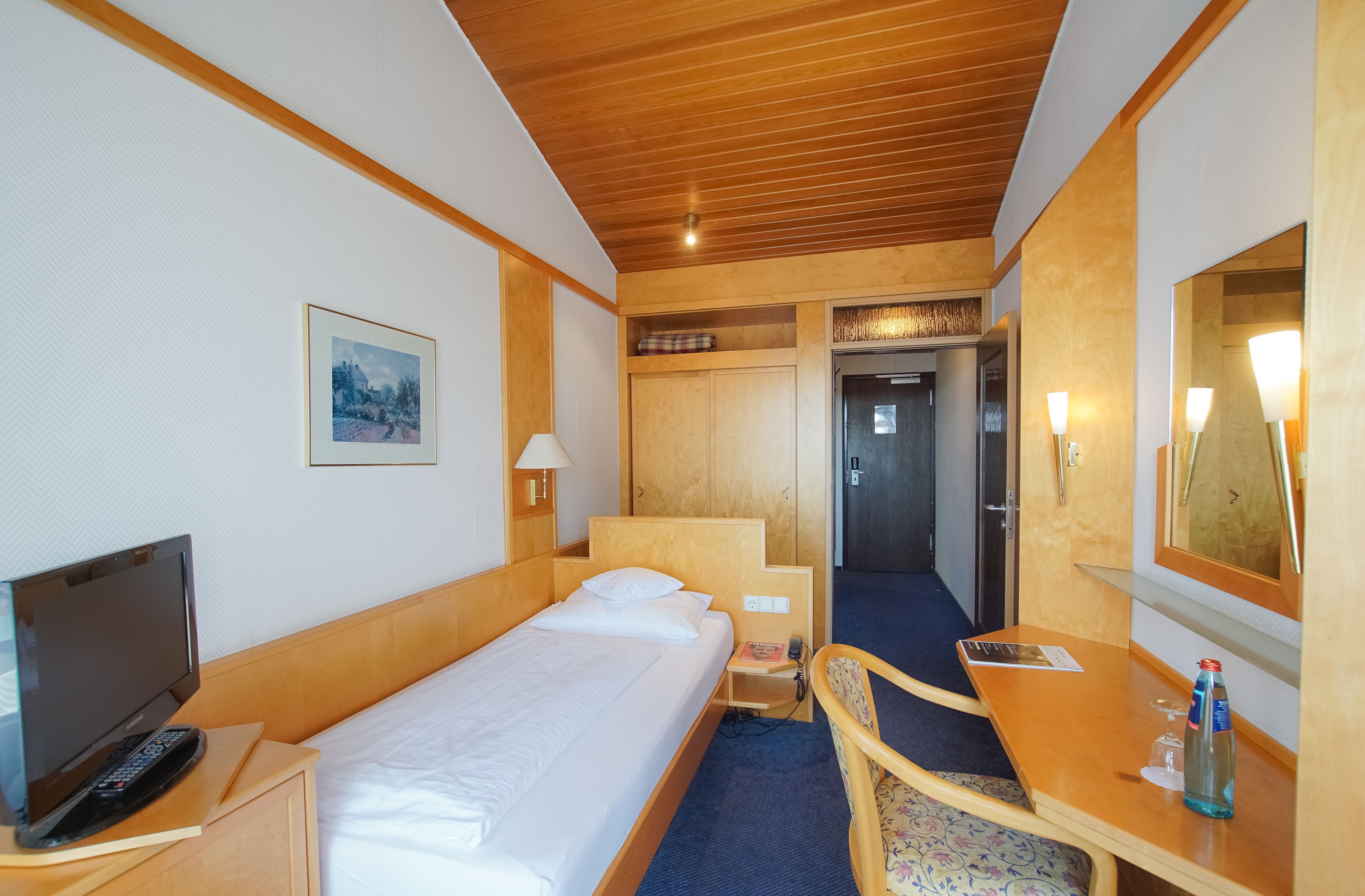 A hotel room with a bed, bedside table with a lamp, a desk with a chair, a television, and a mirror.