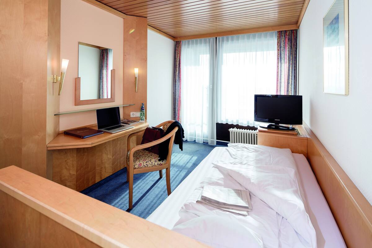 A hotel room with a bed, a desk with a laptop, a chair, a television, and a window with curtains.