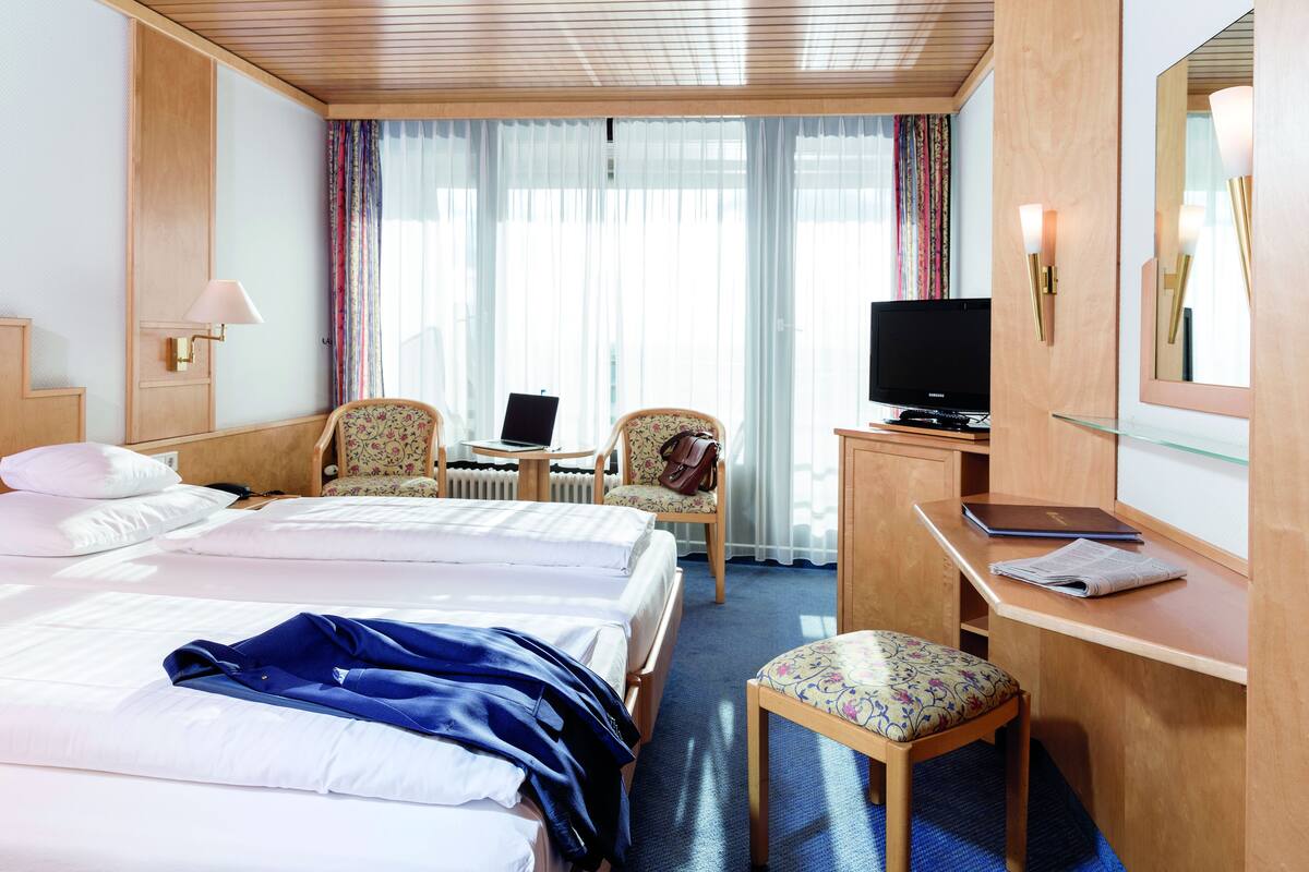 A hotel room with two beds, a desk with a computer, a chair, a television, and a large window with curtains.